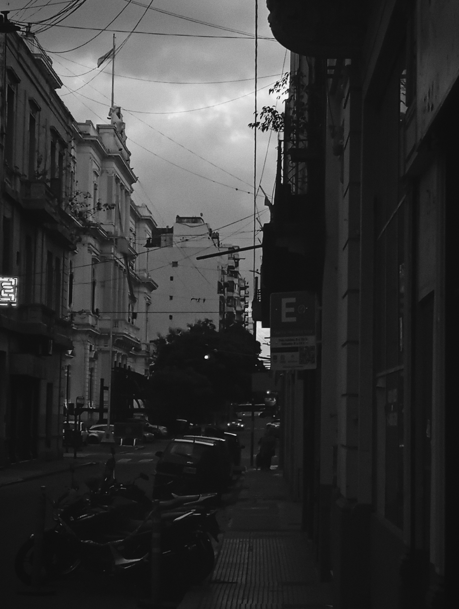 This black and white photograph shows a street scene in Buenos Aires, Argentina. A narrow street is flanked by buildings on either side. On the left, the building is a light-colored, older structure with ornate architectural details, including a flag flying from the top. On the right, the building is darker and more modern with what appears to be storefront windows. There is a parking sign with the letter "E" on it affixed to the building on the right side of the road. Several cars and motorcycles are parked along the street. Power lines crisscross above. The sky is overcast. A person is walking down the sidewalk.