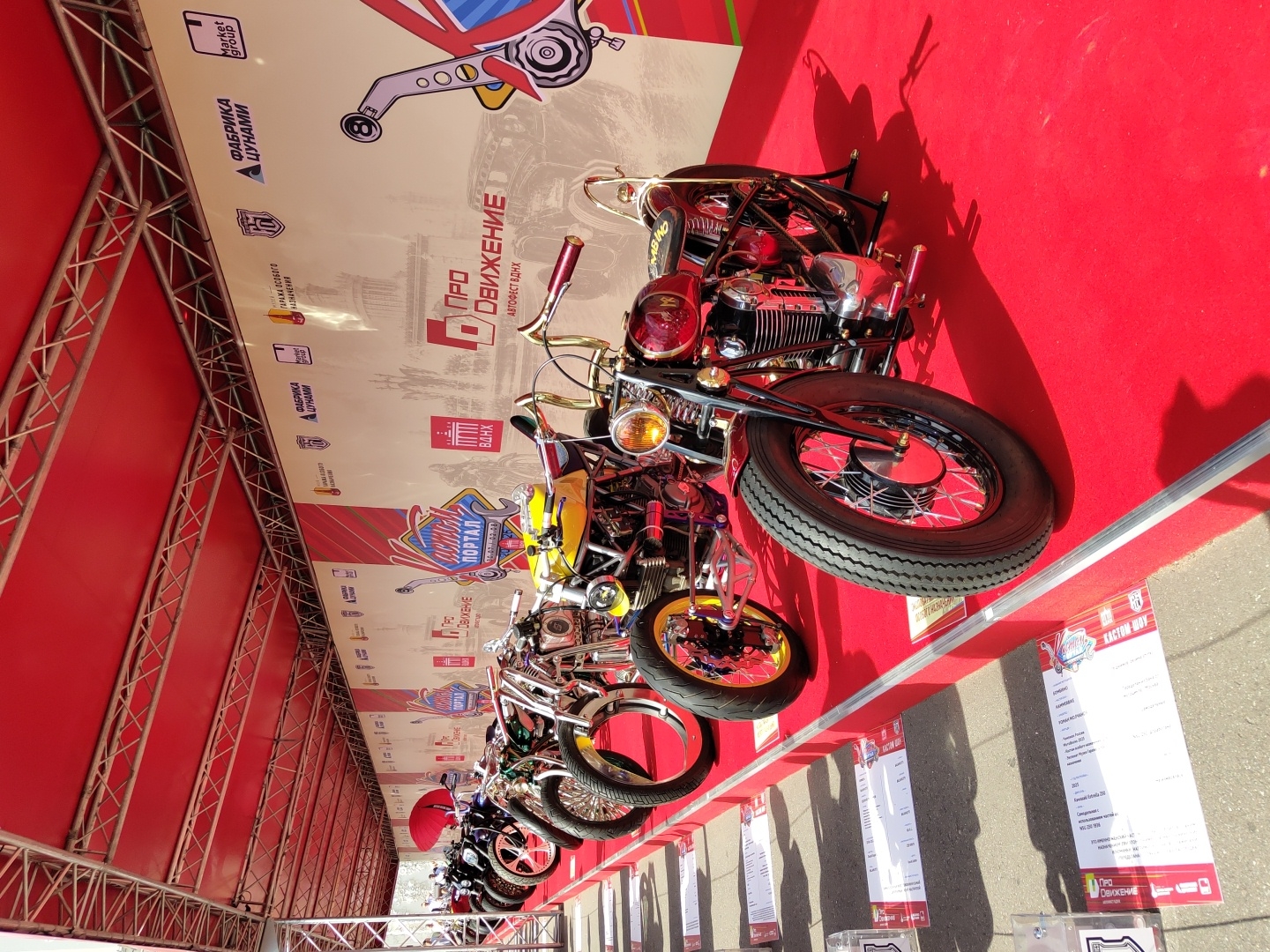 This image depicts an outdoor exhibition of custom motorcycles, set up on a red carpet under a red canopy structure. The exhibition appears to be part of a larger event, as indicated by the banners and signage. The banners in the background feature Cyrillic text, including "ПРО ДВИЖЕНИЕ" (Pro Dvizhenie) and "ВДНХ" (VDNKh). VDNKh is a well-known exhibition center in Moscow, Russia.
The foreground of the image showcases several meticulously detailed motorcycles. The motorcycle closest to the viewer is a red custom bike with chrome accents, a large spoked front wheel with a vintage tire, and a visible engine. To its left, a bright yellow custom motorcycle with similar spoked wheels and a more compact frame is displayed. Further to the left, a row of black motorcycles is partially visible, suggesting a diverse collection of custom bikes.
Several white information placards are placed on the red carpet in front of the motorcycles, likely providing details about each vehicle. The overall environment suggests a sunny day, with shadows cast by the motorcycles and the canopy structure. The background banner also includes logos of sponsors, such as "ФАБРИКА ЦУНАМИ" (Fabrika Tsunamii) and "Market group". The presence of VDNKh, a prominent Moscow landmark, confirms the location.