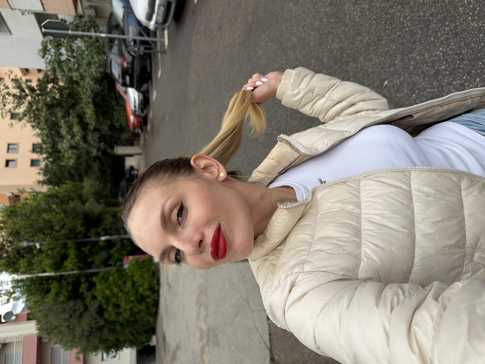 A woman with fair skin and red lipstick poses outside. She has blonde hair pulled back in a ponytail, wearing a white t-shirt and a light beige puffer jacket. She is holding the end of her ponytail in her hand. The background shows a street with parked cars and buildings. The location is in Budva, Montenegro.