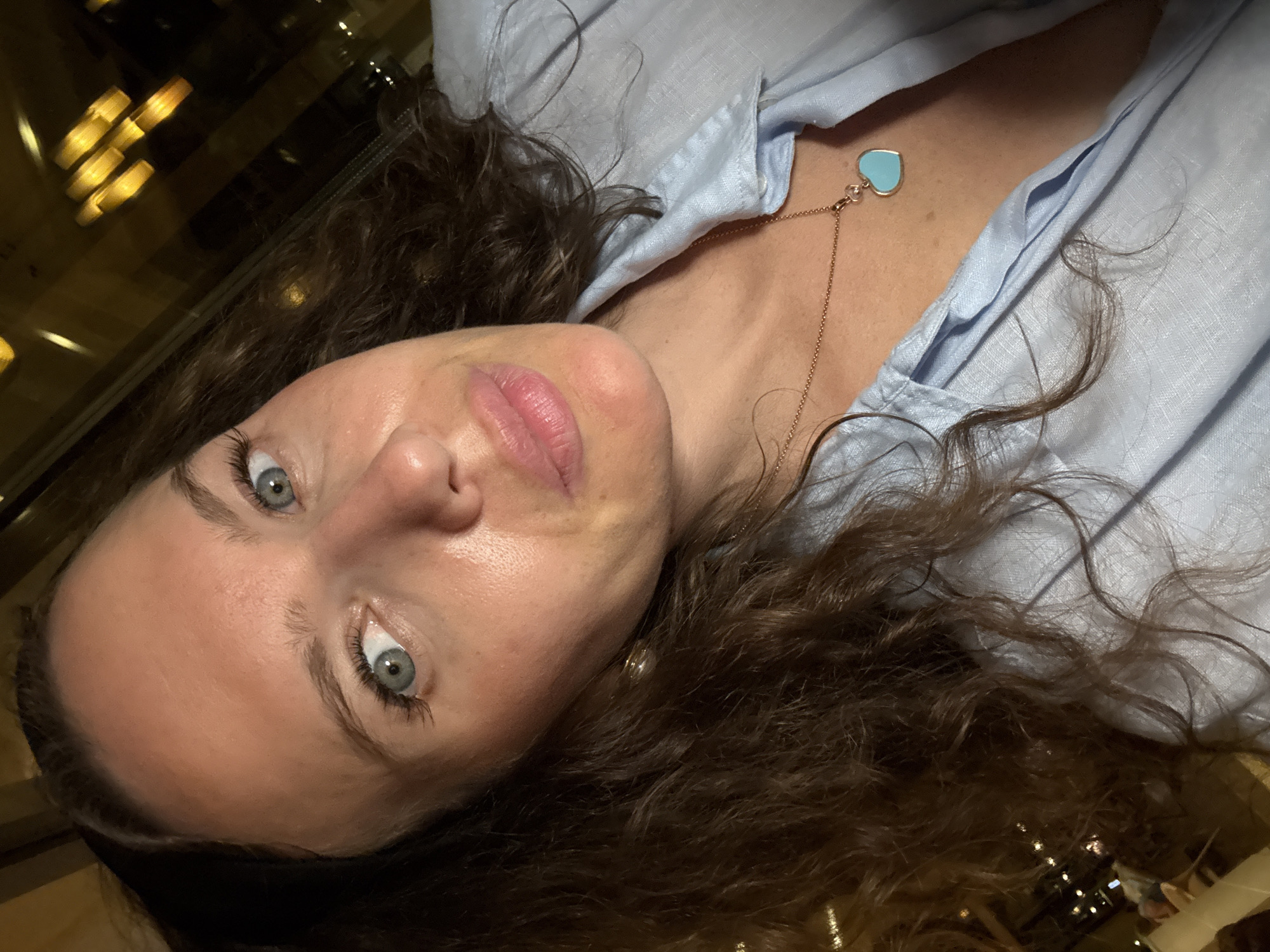 A woman with curly brown hair, blue eyes, and fair skin takes a selfie. She is wearing a light blue linen shirt and a rose gold necklace with a turquoise heart pendant. The background appears to be an indoor setting with blurred yellow lights visible through a window, suggesting it might be nighttime. There is no identifiable information about the location being Dubai, United Arab Emirates.
