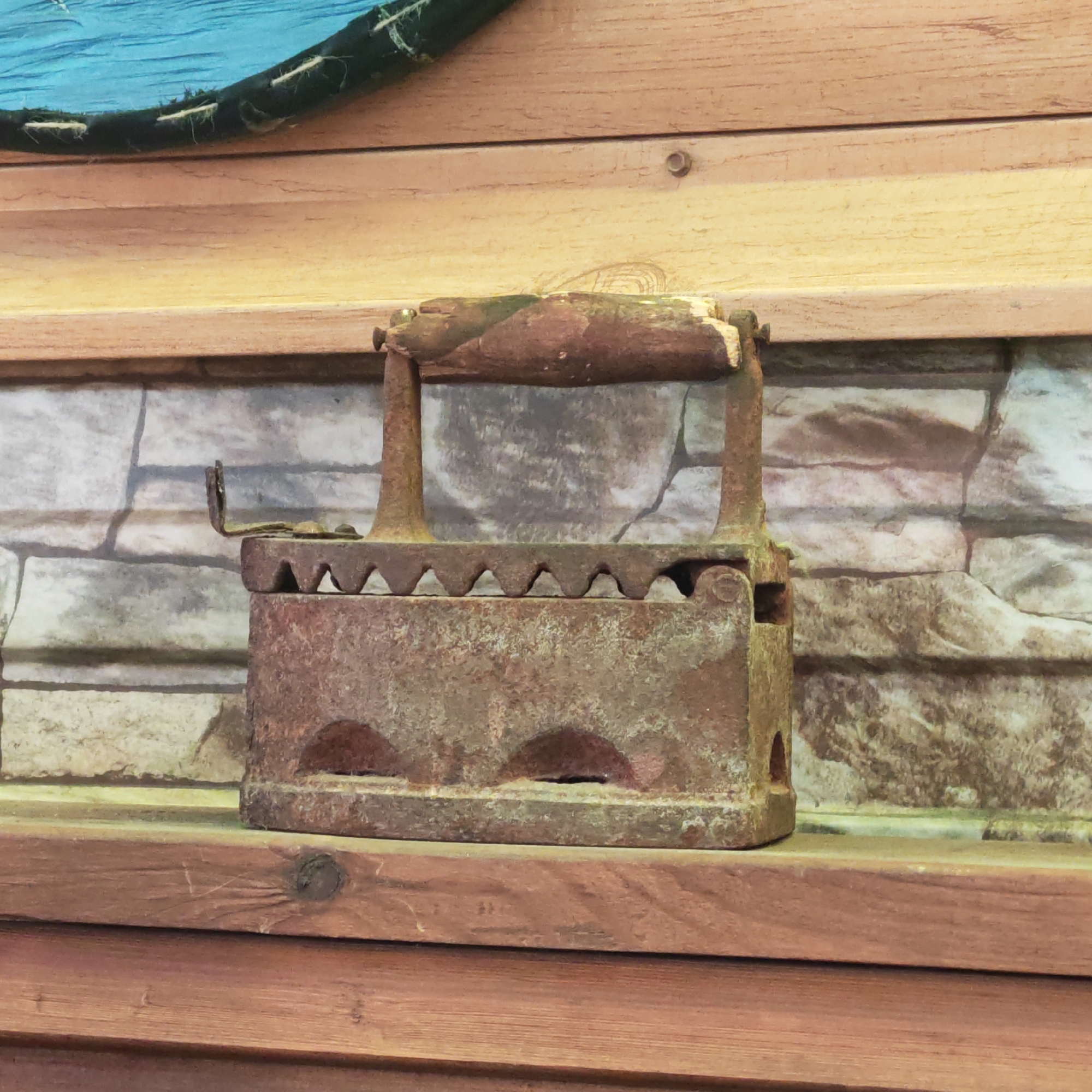A rusted antique iron sits on a wooden shelf against a wall with a faux brick pattern. The iron is heavily corroded, with a dark, reddish-brown color and visible pitting. Its handle is also rusted and appears to be made of wood and metal. The shelf is made of multiple horizontal wooden planks, with a reddish-brown hue. The wall behind the shelf has a printed brick design. The scene is well-lit. The image was taken in Rostov, Russia.