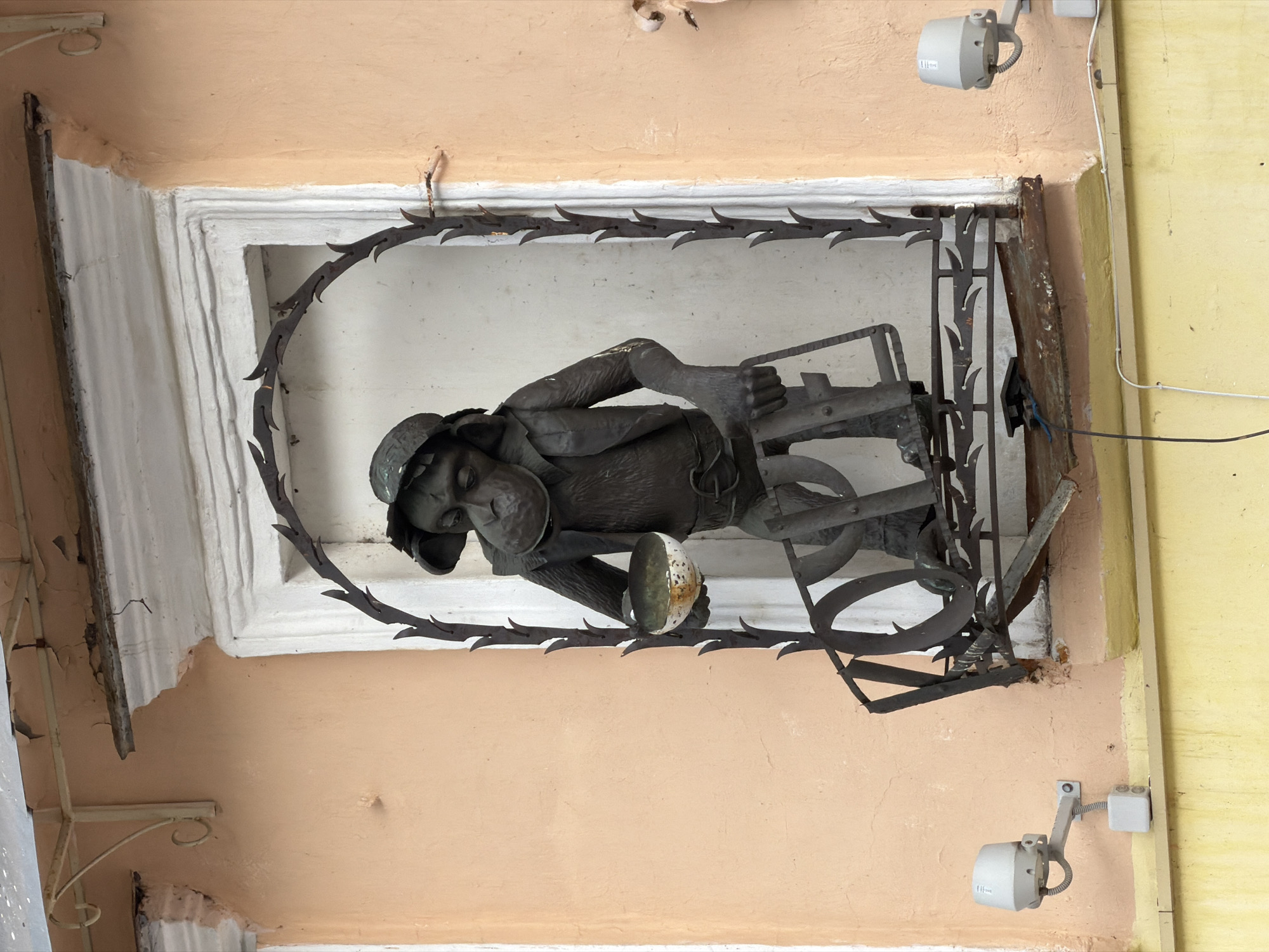 The image shows a bronze sculpture of a monkey in a window niche on the facade of a building in Nizhny Novgorod, Russia. The monkey is depicted wearing a hat, a vest, and trousers, and is holding a bowl in its left hand. The sculpture is mounted on a decorative metal railing that extends across the window. The building's facade is painted in a peach color, and the window frame is white. There are two security cameras mounted on the wall below the window, one on each side. The overall scene suggests an outdoor urban environment, possibly a street or a public square. The sculpture's detailed craftsmanship and its placement in the window niche indicate it's a decorative element of the building.