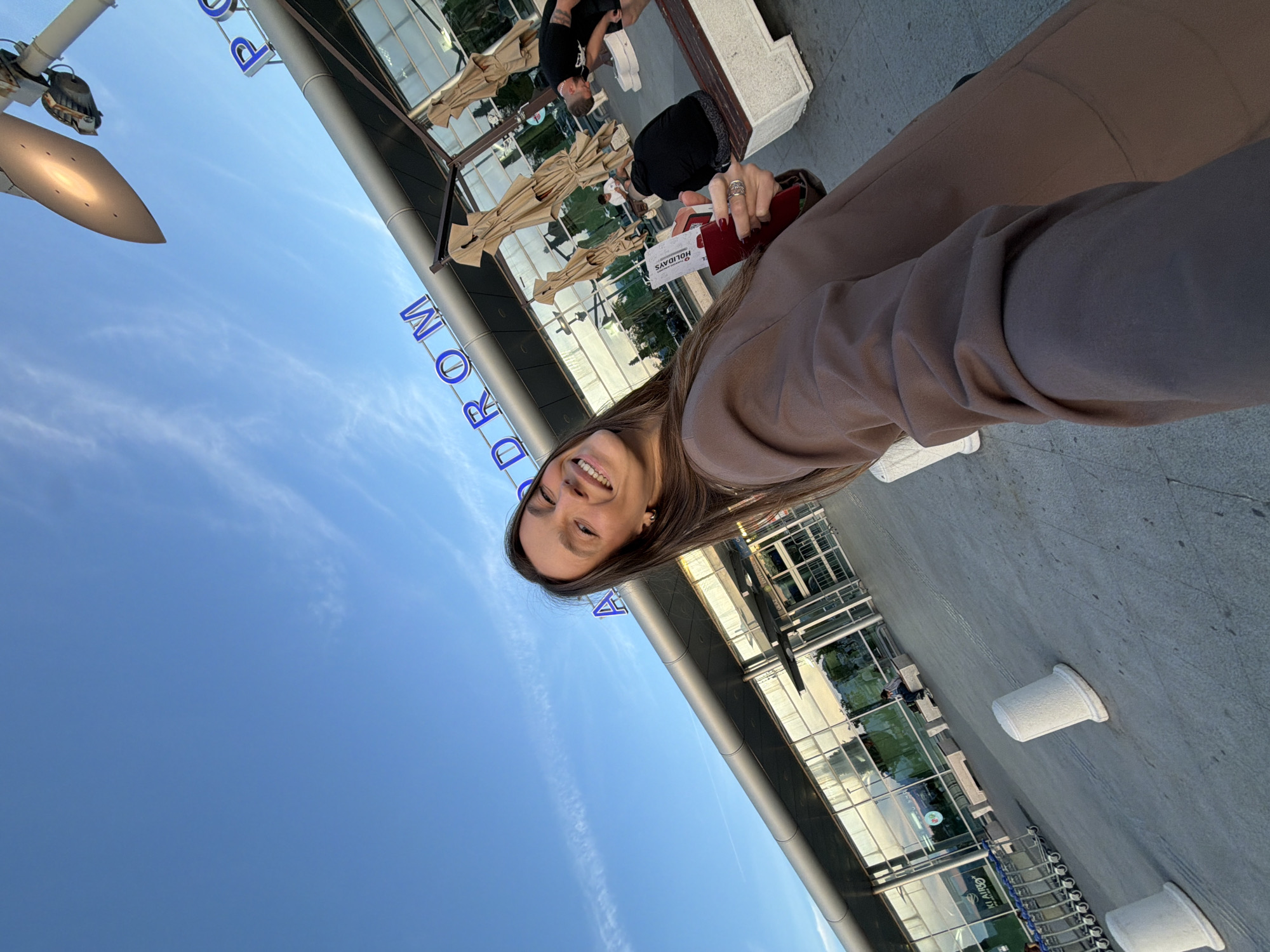 A woman is taking a selfie outside the Podgorica Airport in Golubovci, Montenegro. She is holding a boarding pass and a red passport. Behind her is the airport building with glass windows and doors, and blue signage that reads "PODROM". There are people sitting at tables with umbrellas in front of the building. The sky is blue with some clouds. There are also white cylindrical objects lined on the ground.