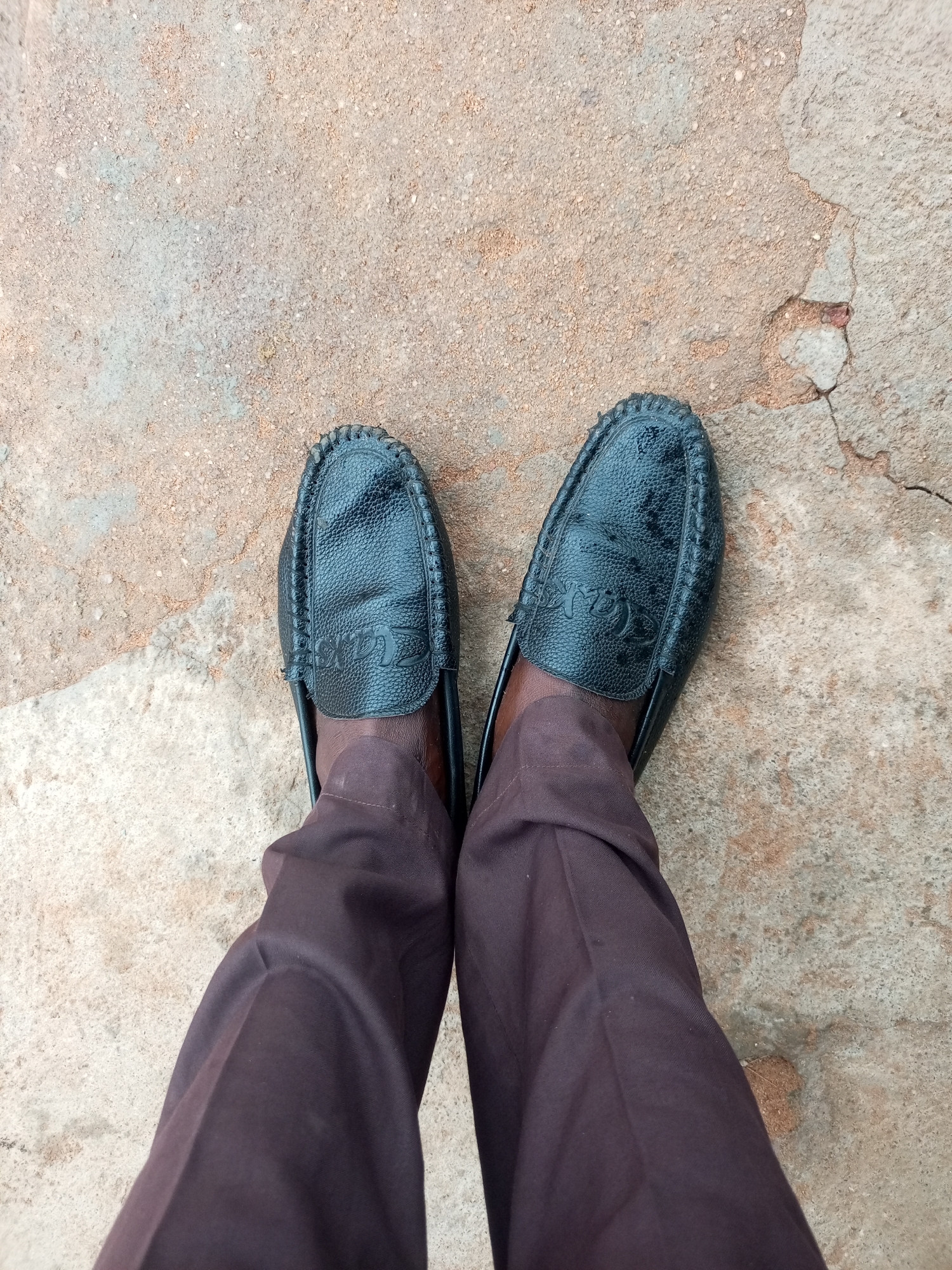 The image shows a pair of black leather moccasin-style shoes with a textured surface and decorative stitching around the edges. The shoes have a subtle logo embossed on the vamp. The person wearing the shoes is also wearing dark brown trousers. The shoes and trousers are positioned on a cracked, concrete surface. The concrete is a light brown color with some lighter patches and visible damage, suggesting an outdoor environment. The perspective is from above, looking down at the feet. No specific location in Tempure, Nigeria, can be determined from the image.