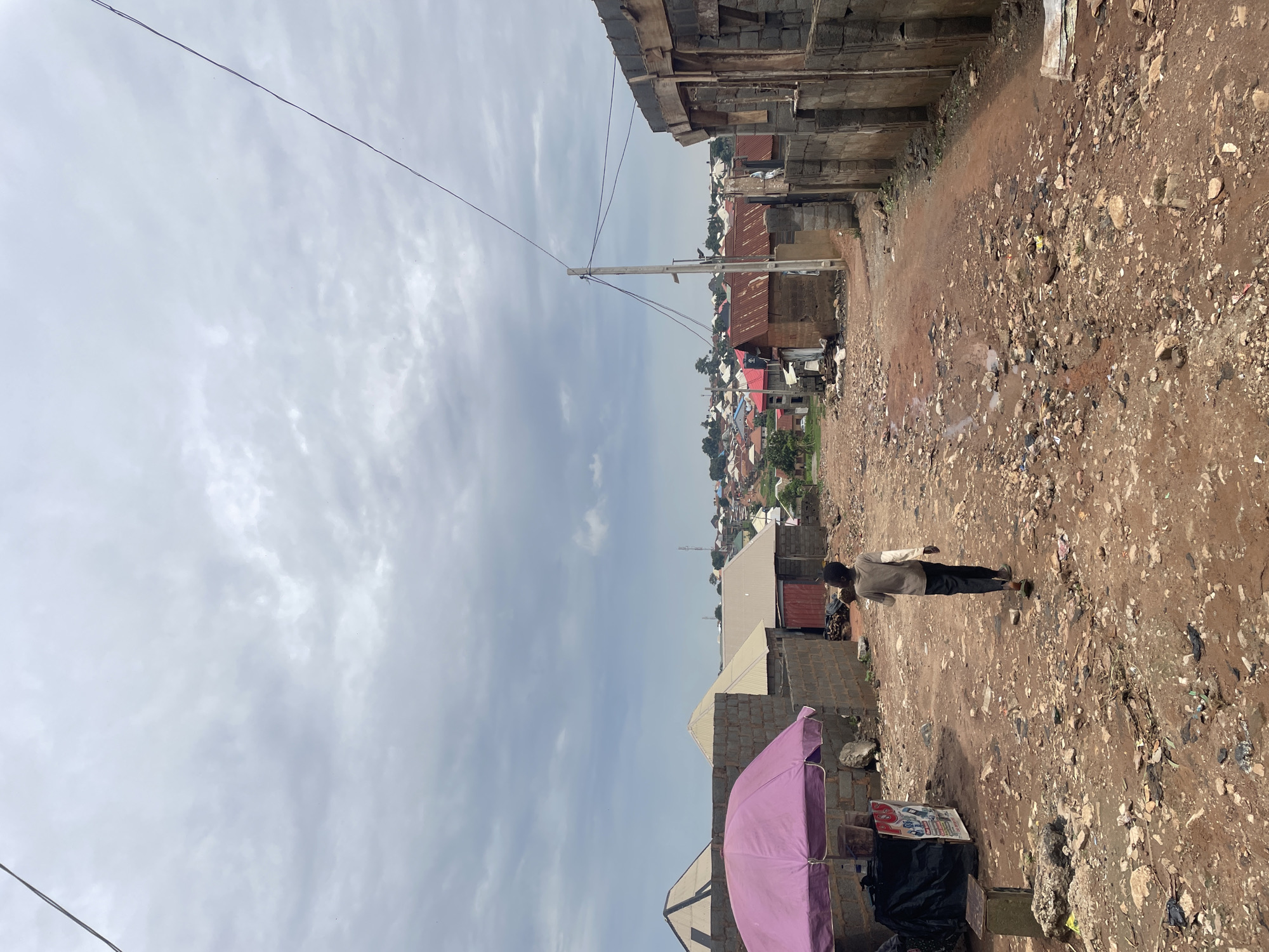 The image depicts a dirt road in Kurunduma, Nigeria, under a cloudy sky. A young boy, wearing a light-colored shirt and dark pants, walks away from the camera down the road. He is carrying something over his left shoulder. To the left of the road, a pink umbrella shades a small stall with a black tarp and a sign. The road is unpaved and covered in rocks, dirt, and some scattered debris. The surrounding buildings are constructed from concrete blocks, some unfinished. In the background, a densely populated area with various buildings, many with corrugated metal roofs, extends into the distance. Power lines are visible overhead.
