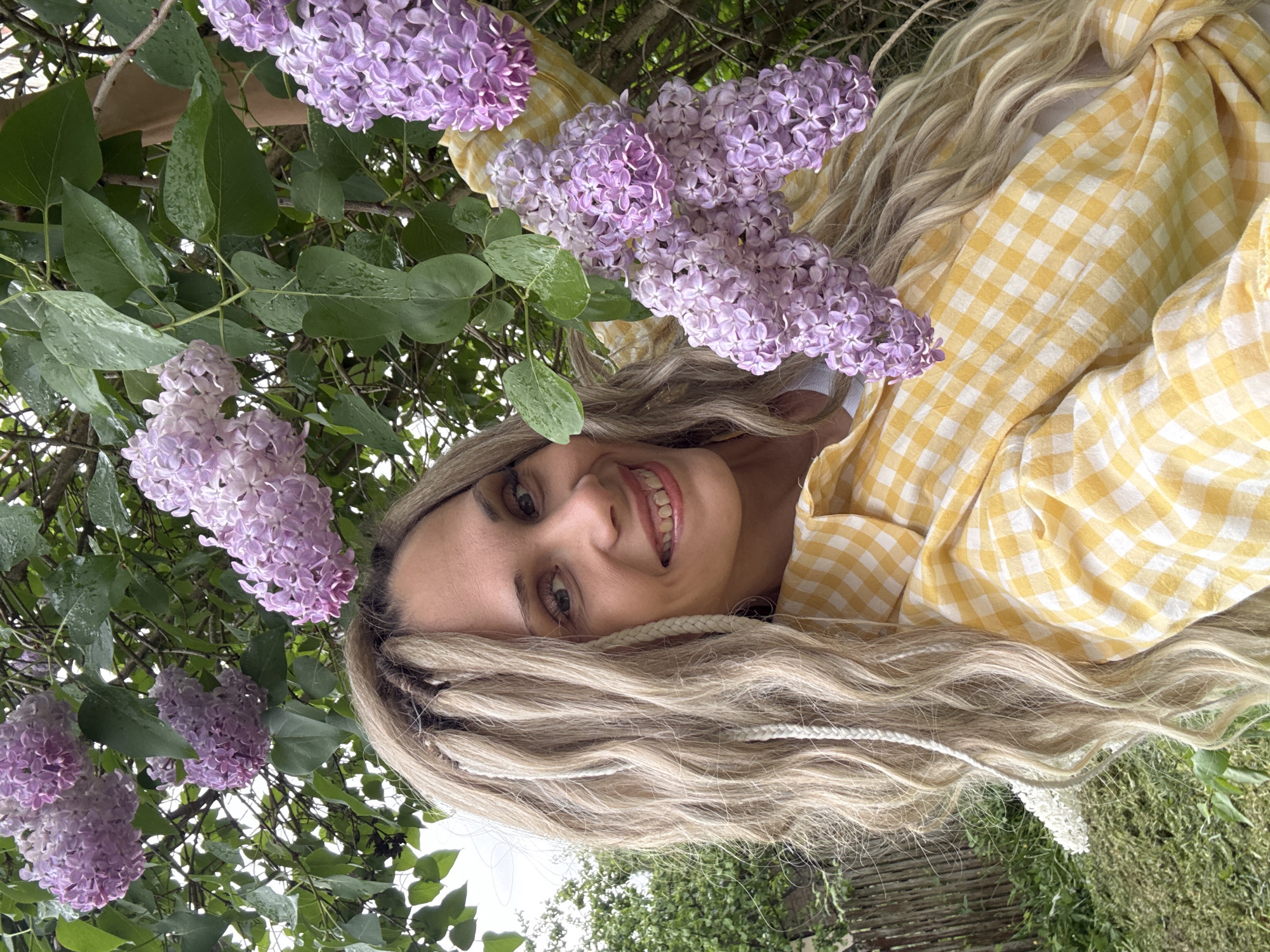 The image shows a woman with long, blonde hair and a yellow and white checkered shirt lying down outdoors. She is surrounded by lilac flowers and green leaves, possibly in a garden or park. The background includes glimpses of vegetation and possibly a wooden fence.