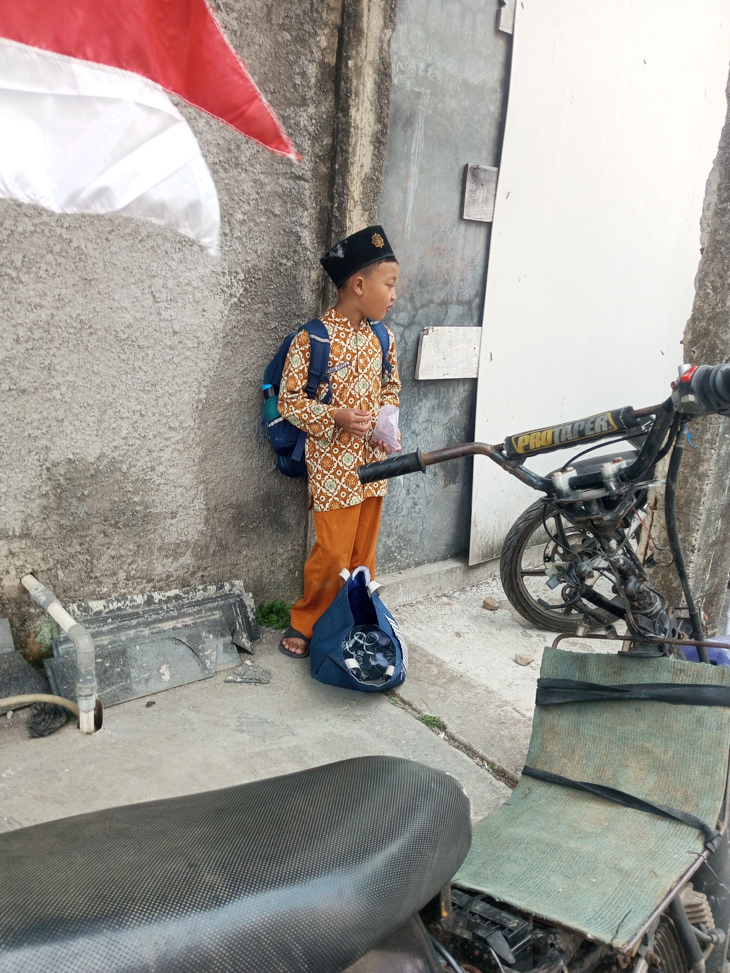 The image depicts a young boy standing on a concrete surface next to a rough, textured wall. He is wearing a patterned shirt, orange pants, and a black peci hat. A blue backpack is slung over his shoulders, and he is holding a small white object in his right hand. In front of him, a blue bag lies on the ground. To his right, a motorcycle is partially visible, with its handlebars and front wheel showing. A portion of a red and white flag is draped from the upper left corner of the frame. The environment appears to be an outdoor urban setting, possibly an alleyway or side street. The wall exhibits signs of age and weathering, with patches of discoloration and debris scattered on the ground. The motorcycle seems to be a custom or modified bike, with visible wiring and a stripped-down frame. The scene is illuminated by natural daylight, casting soft shadows. The overall impression is one of everyday life in a locality, potentially in the city of Kalibeber, Indonesia, given the context provided.
