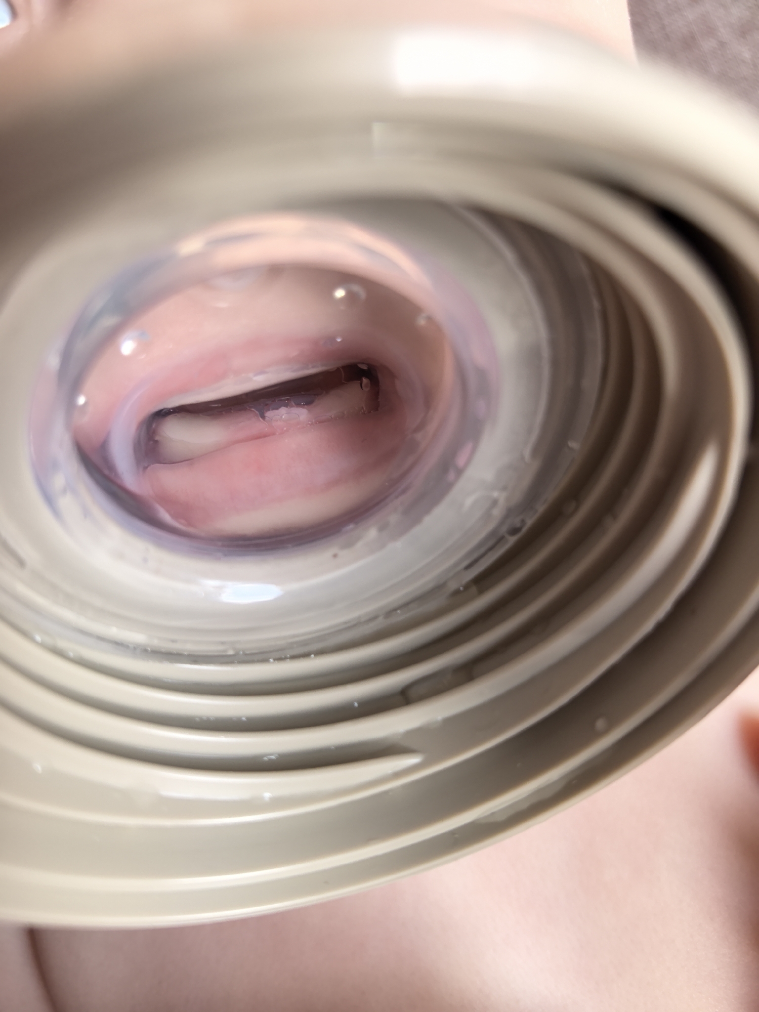 A close-up, slightly out-of-focus image shows a person's mouth viewed through a circular opening, possibly a bottle or container top. The mouth is open, revealing the upper and lower lips, tongue, and some teeth. There's a clear, slightly purple-tinted liquid or substance in the opening and around the mouth. The surrounding object is beige or light brown with concentric ridges, likely made of plastic. No specific location in Фролы, Russia, can be determined from the image. There is no specific information available about people, subjects, objects, actions, interactions, or the environment.