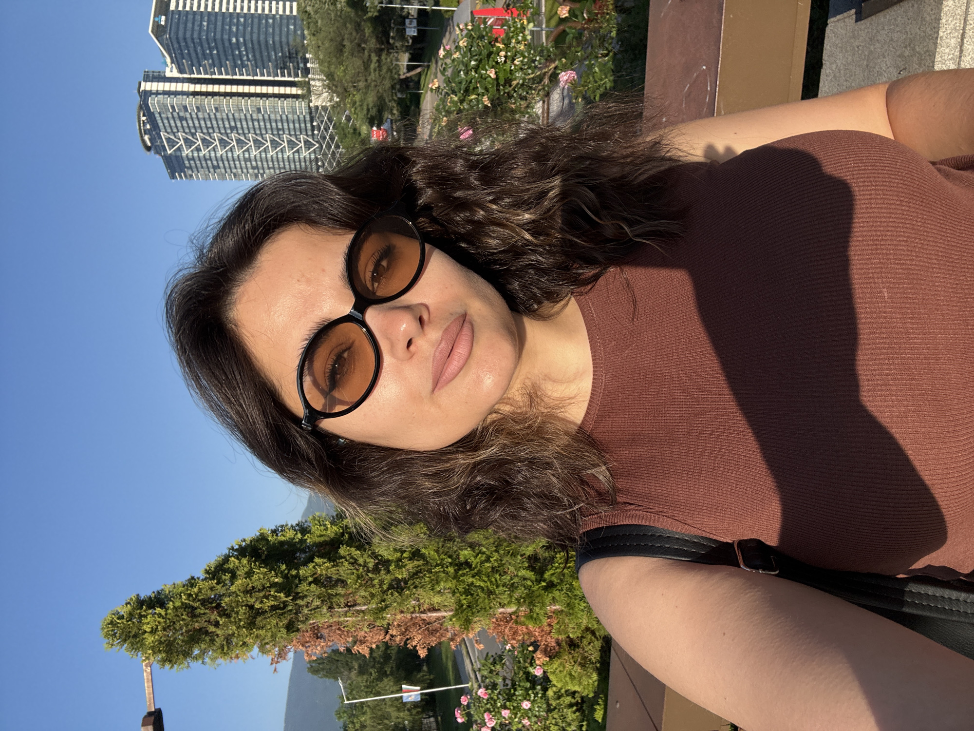 A woman with dark, curly hair and sunglasses is taking a selfie outdoors. She is wearing a brown, ribbed tank top. Her right arm is visible, and she is wearing a black backpack strap over her shoulder. In the background, there are trees, flowers, and modern skyscrapers. The sky is clear blue. The lighting suggests it is late afternoon.
