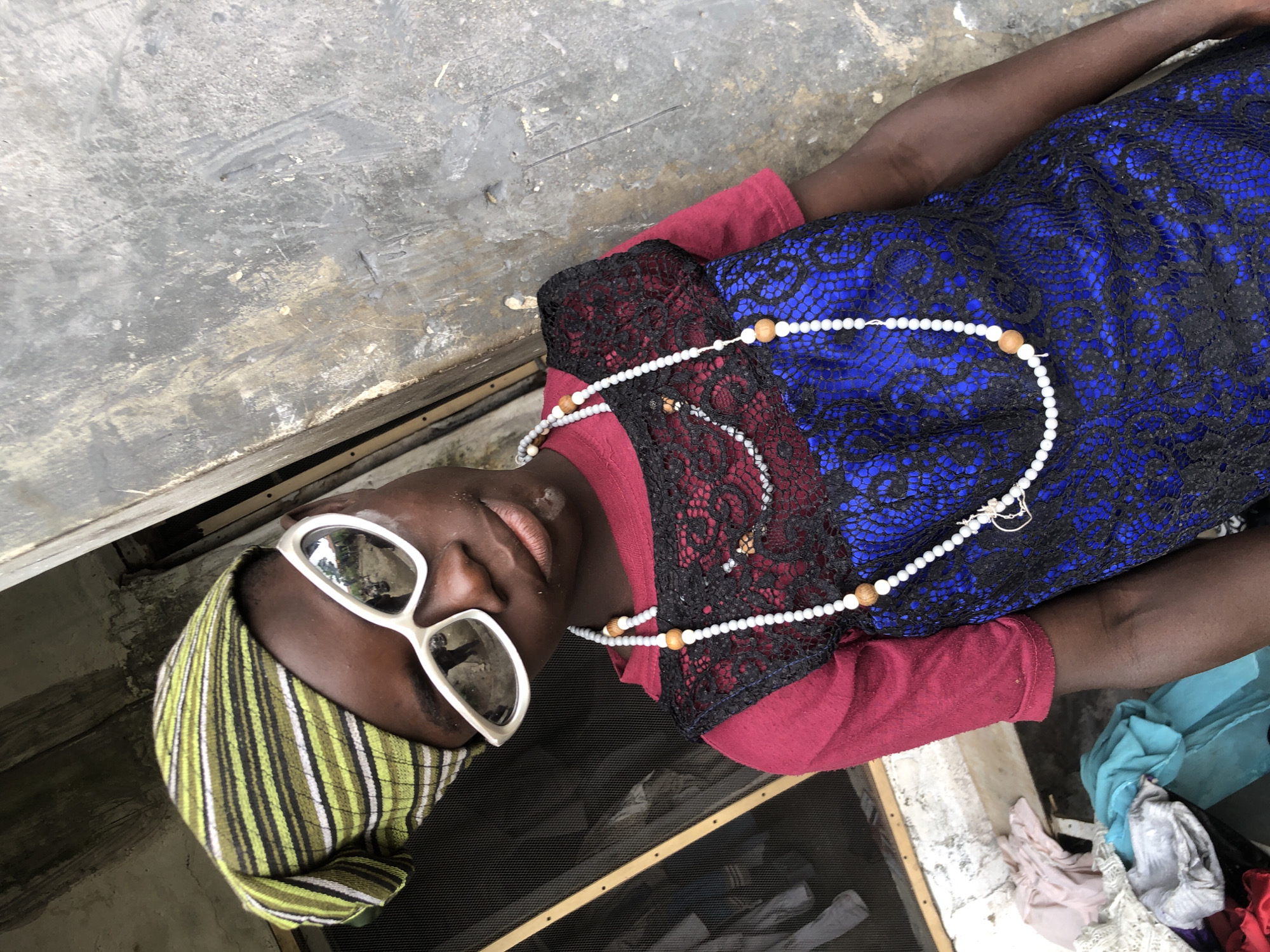 The image is a medium shot of a young person, possibly a child, in Ado, Nigeria. They are wearing a green and white striped headwrap, white-rimmed sunglasses, a maroon shirt, and a blue dress with black lace overlay. A long string of white and wooden beads is draped around their neck. The background consists of a weathered concrete wall and a screened window. Piles of colorful fabric are visible behind the screen. The person is looking towards the camera with a neutral expression.