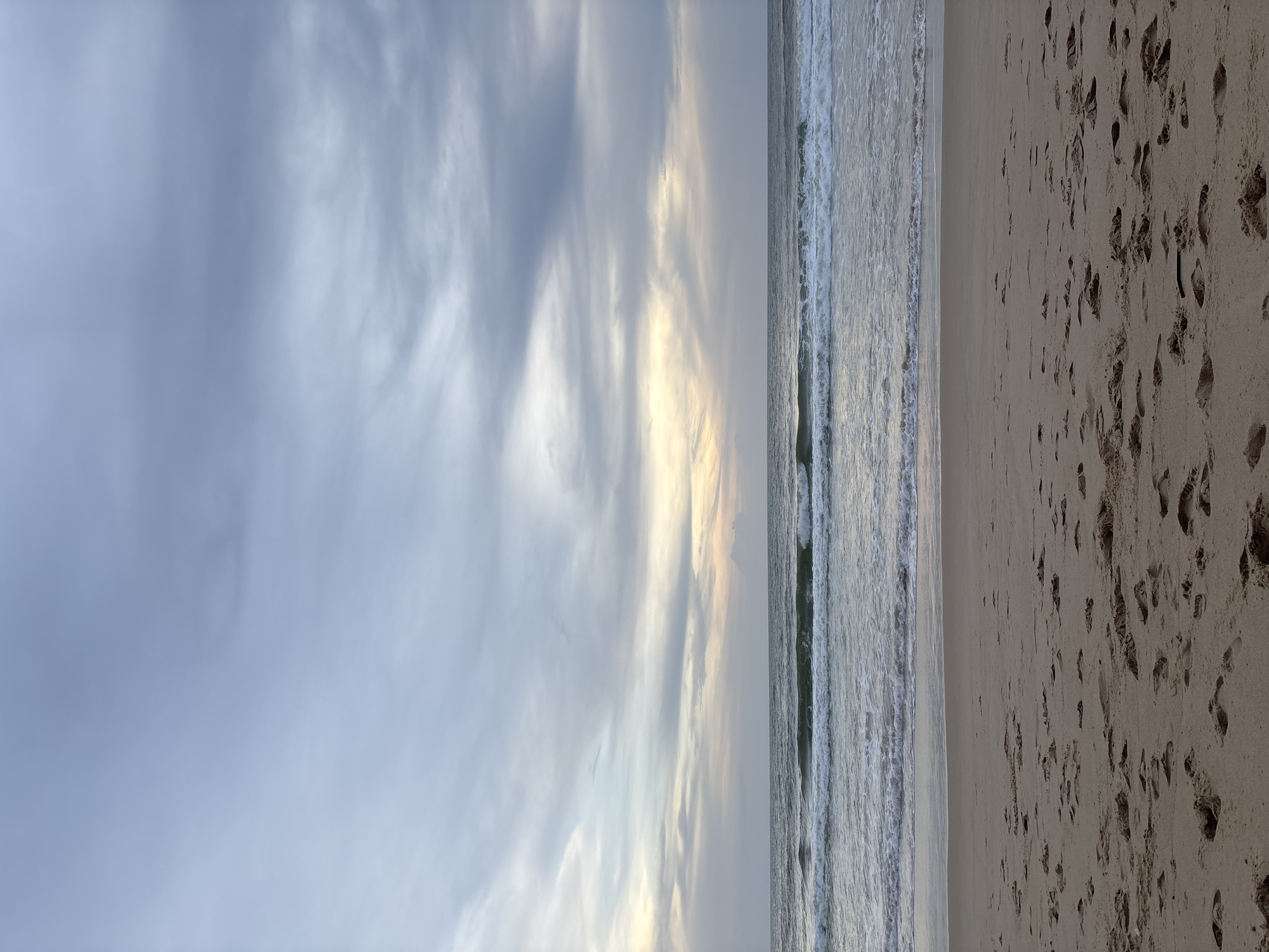 This is a landscape image of a beach scene in Choeng Thale, Thailand. The sky is mostly cloudy with some patches of lighter coloration, possibly indicating the presence of sunlight. A sandy beach occupies the right portion of the image, marked with numerous footprints. The sea is positioned in the middle, with small waves breaking onto the shore.