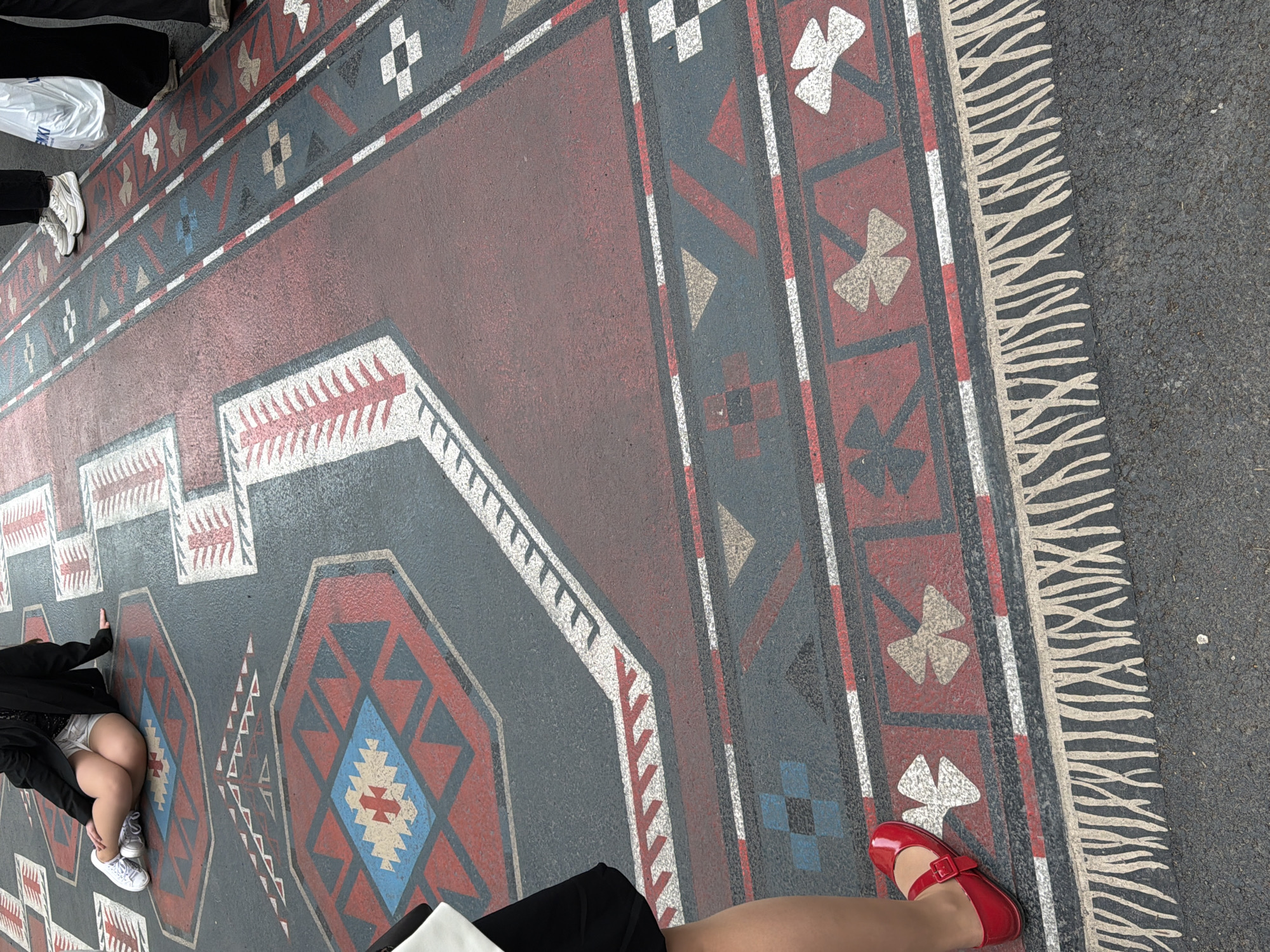 The image shows a ground-level view of a large, patterned area designed to resemble a traditional Armenian rug. The "rug" is painted onto a paved surface and features geometric designs in red, blue, black, and white. The "fringe" of the rug is also painted.
Several people are present in the image, partially visible. One person is wearing a black jacket and shorts and white sneakers. Another person is wearing black pants and a white top, and red Mary Jane shoes.
The overall setting appears to be a public space, likely a street or square in Yerevan, Armenia, given the reference to an Armenian rug design. The pavement outside of the rug design is asphalt.