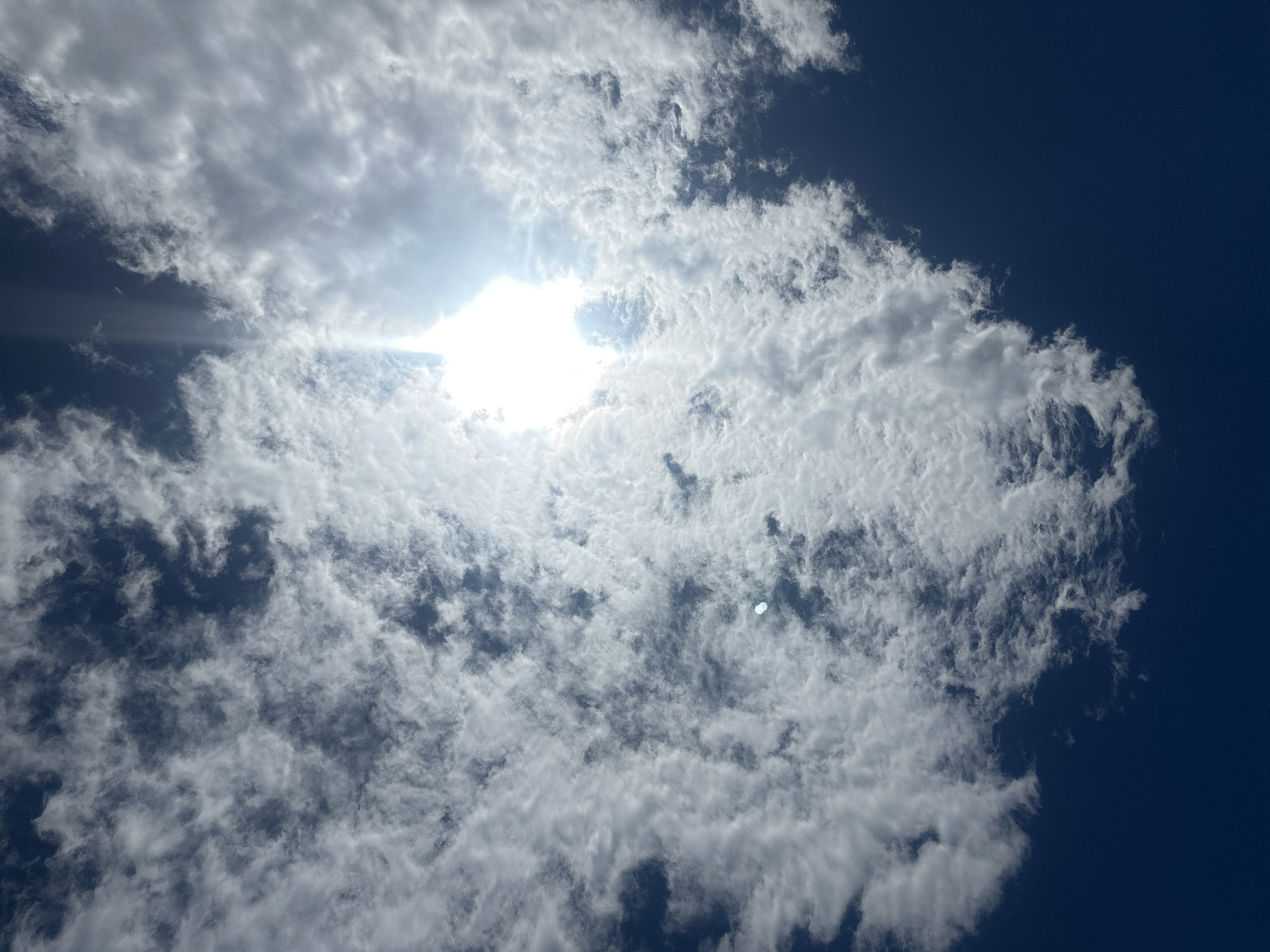 A daytime, upward-facing shot shows a cloudy sky, likely in Sant Feliu de Guíxols, Spain. A dense cluster of white clouds occupies the majority of the frame, with the sun shining brightly through a gap in the clouds. The sky visible around the clouds is a deep blue. No people, subjects, or specific objects beyond the sky and clouds are identifiable. No actions or interactions are visible in the frame.