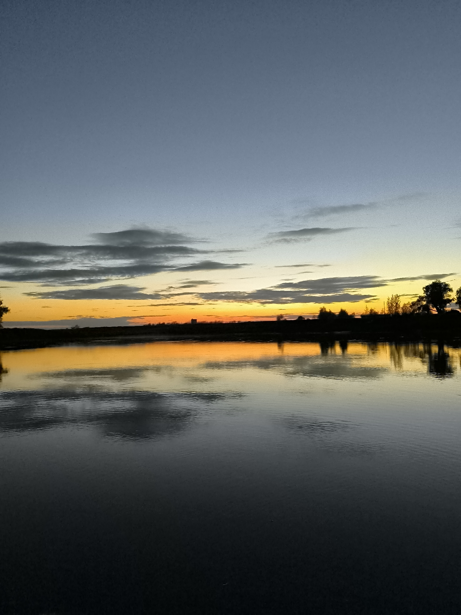 This is a photograph of a serene lake at dusk, capturing the transition from day to night in Zagotzerno, Russia. The sky is a gradient of soft blues and grays at the top, transitioning to vibrant oranges and yellows near the horizon, indicating the setting sun. Wispy clouds are scattered across the sky, reflecting the warm hues of the sunset.

The horizon is lined with a dark silhouette of trees and what appears to be a few distant buildings. The lake in the foreground is calm and still, creating a perfect mirror reflection of the sky and the treeline. The water's surface is dark, with subtle ripples disrupting the otherwise smooth reflection. The overall mood of the image is peaceful and tranquil, evoking the quiet beauty of nature at the end of the day. There are no people visible in the image, and the environment is natural and undeveloped except for the distant structures.