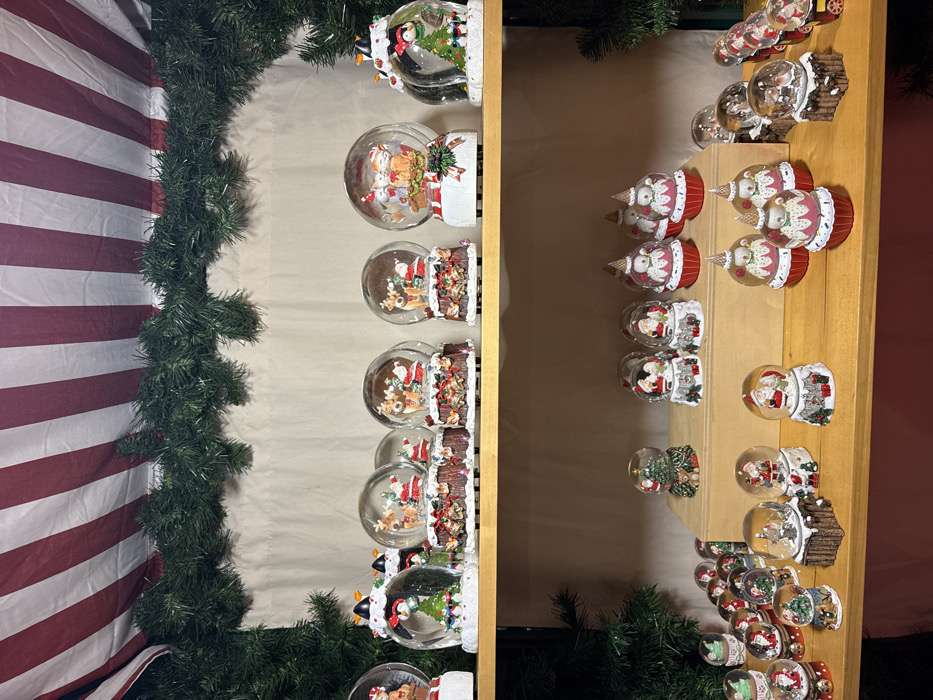 A shelf displays a collection of snow globes, mostly Christmas-themed. Many feature Santa Claus with reindeer, snowmen, or penguins inside. The bases are decorated in holiday styles, including festive red and white stripes, log cabins, and icy designs. A garland of green pine needles frames the left side of the shelf, while a striped red and white fabric hangs nearby.