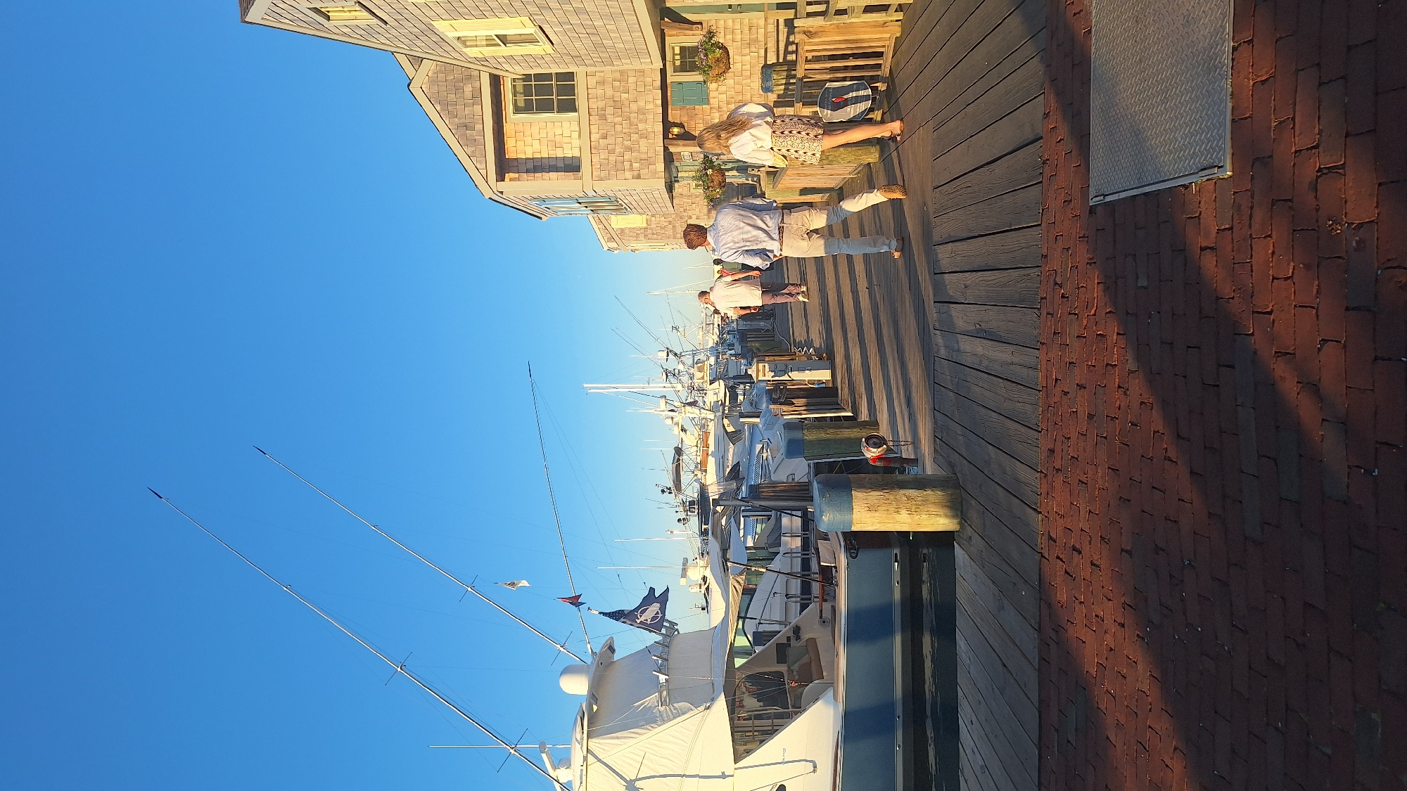 The image depicts a waterfront scene in Nantucket, United States of America, likely in the historic harbor area. The foreground features a brick walkway transitioning to a wooden pier. Along the pier, several large white yachts are moored, their tall masts and rigging silhouetted against a clear blue sky. The yachts are docked side-by-side, creating a sense of a bustling marina.
On the pier, several people are walking. In the mid-ground, a couple is walking away from the camera. The man is wearing a light blue button-down shirt and light-colored pants, while the woman is in a white shirt and a patterned skirt. Further down the pier, another person in a light-colored shirt and pants is walking towards the boats.
To the right of the pier, there are buildings with a distinctive shingle-style architecture, characteristic of Nantucket. These buildings appear to be shops or restaurants, with windows and some decorative elements like hanging flower baskets. The setting sun casts long shadows across the pier, indicating it is late afternoon or early evening. The overall atmosphere is one of a pleasant, upscale maritime setting.