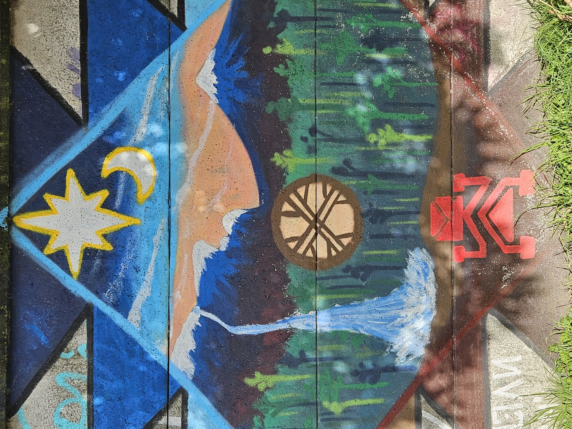 The image shows a section of a concrete wall adorned with colorful graffiti. The graffiti includes a blue diamond with a white star and crescent moon outlined in yellow, a peach-colored shape with white accents, a brown circle containing a stylized tree-like design, green and blue streaks resembling foliage and water, and a red geometric symbol possibly representing the letter "K". The concrete sections are separated by vertical lines. Some grass is visible along the right edge of the image. The location is Osorno, Chile.