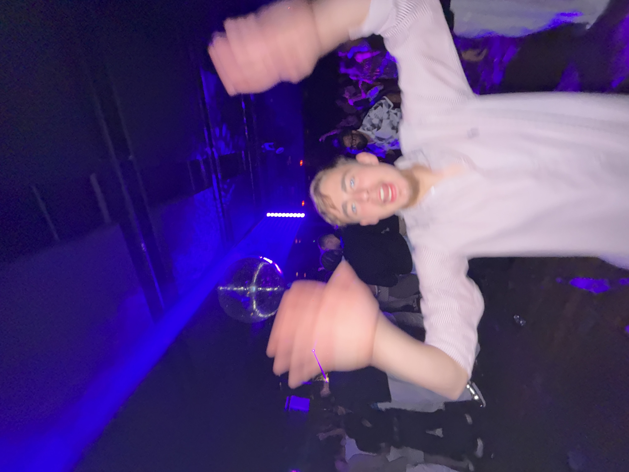 The image appears to be a blurry photograph taken inside a nightclub, likely in Lisbon, Portugal, given the prompt's context. A young man with blond hair and blue eyes is the central subject, holding his hands up near the camera, obscuring the view. The scene is dimly lit with blue and purple hues dominating the color palette. A disco ball is visible in the background. There are multiple people in the background, but their features are indistinct due to the low light and blur. Some individuals appear to be wearing face masks. The overall impression is of a lively and crowded nightlife setting.