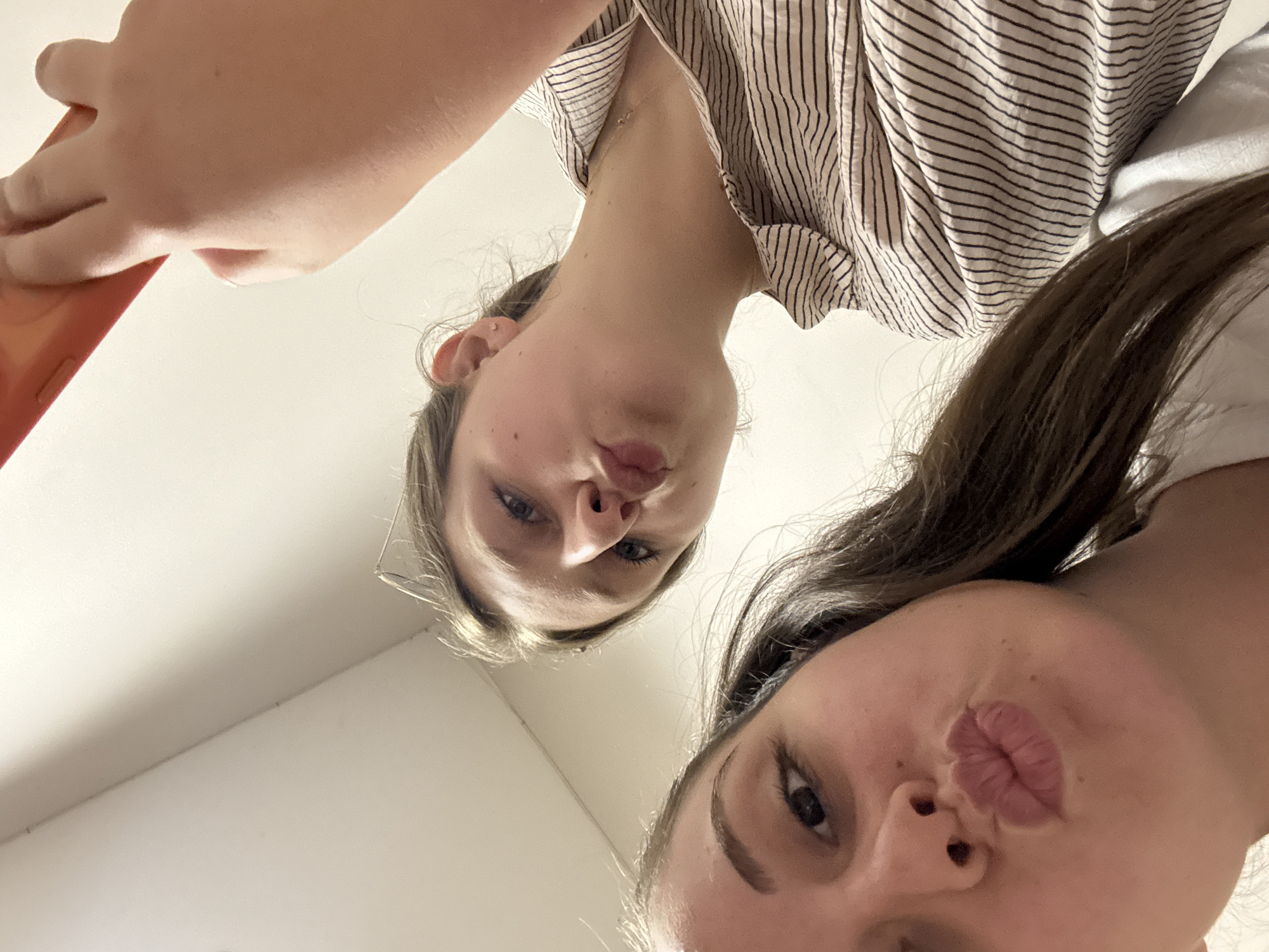 Two young females, appearing to be teenagers, are posing for a selfie taken from a low angle. The female in the foreground is on the left, with brown hair. The female in the background is on the right, with lighter brown hair. Both are puckering their lips. The female on the right is wearing a striped shirt and holding an orange object, possibly a phone case. The background appears to be an indoor setting with a white ceiling and wall. There is no indication of the city of Burgas, Bulgaria in the image.