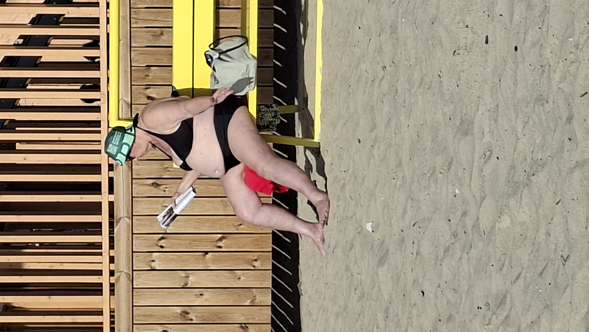 A person is sitting on a yellow bench on a sandy beach. The person is wearing a black bikini and a green and white patterned hat. They are holding a magazine and appear to be reading it. To the right of the person is a light-colored bag. Behind the bench is a wooden fence, and beyond that, a wooden structure. The ground is covered in sand, with some debris visible. The scene appears to be outdoors, possibly a beach resort or public beach. There is no specific indication of the location being in Tallinn, Estonia.