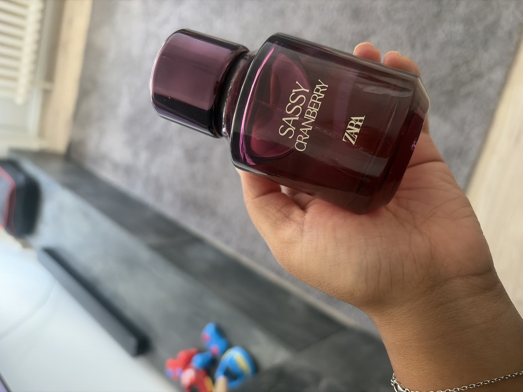 A person's hand holds a dark red glass perfume bottle with a matching dark red cap. The bottle has white text on it that reads "SASSY CRANBERRY" and "ZARA". The person is wearing a silver chain bracelet on their wrist. The background is blurred, but appears to be an indoor setting with a gray carpet and a dark gray, possibly leather, couch or bench. There are some brightly colored toys, red and blue, on the surface in the background. The scene does not provide any information about the city of Duisburg, Germany.