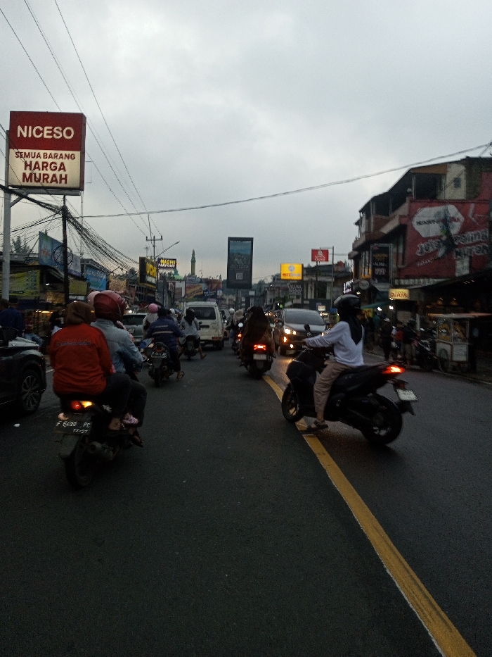 The image captures a busy urban street scene at dusk, likely in the city of Dawathang_Dorjibi_ Kashingtsawa, Bhutan, under an overcast, light gray sky. The asphalt road, appearing damp, features a prominent solid yellow line marking the center or a lane division. The scene is dominated by a dense flow of vehicles, primarily motorcycles, alongside cars and a white van, all moving away from the viewer.
In the foreground, on the left, the rear section of a dark car is partially visible. Behind it, a motorcycle carries two individuals; the driver wears a light blue jacket and a red helmet, while the passenger, positioned behind, wears a red jacket and a dark hijab. The visible license plate on this motorcycle is "F 8430 PC". In the foreground on the right, a dark scooter is ridden by a single individual wearing a white long-sleeved top, dark pants, and a dark hijab, with their left foot near the yellow lane marker. Numerous other motorcycles with riders, some wearing helmets and hijabs, are seen further down the road, interspersed with cars, including a dark sedan with illuminated headlights.
Both sides of the street are lined with multi-story commercial buildings featuring various shopfronts and prominent signage. On the left, a large red billboard displays "NICESO" at the top, with "SEMUA BARANG HARGA MURAH" (Indonesian for "ALL ITEMS CHEAP PRICE") underneath. An intricate network of overhead power lines and cables is visible above the street. On the right, other commercial signs are present, including one with text appearing to read "JELAJAH KULINER NUSANTARA" (Indonesian for "EXPLORE ARCHIPELAGO CULINARY"). In the mid-distance, a green dome and a minaret, characteristic of a mosque, are visible rising above the buildings on the left. The overall environment suggests a bustling commercial area with ongoing street activity.