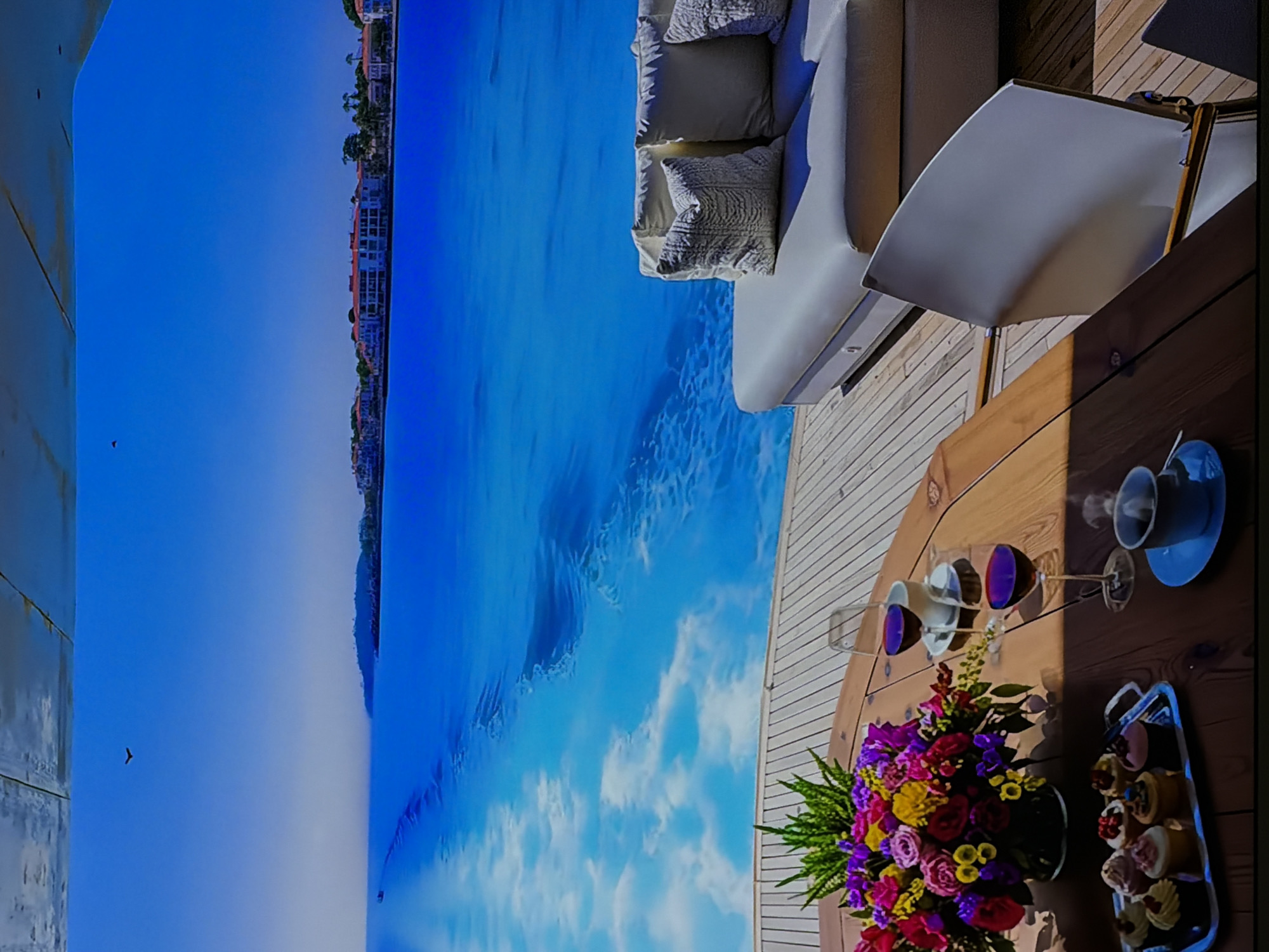 The image displays a serene and luxurious outdoor scene, likely on a yacht or a high-end coastal deck, bathed in bright daylight. In the foreground, a polished wooden deck features a round wooden table. Adorning the table is a vibrant bouquet of fresh flowers in shades of red, pink, yellow, and purple, accompanied by green foliage. Two stemmed wine glasses, containing a dark red liquid, cast distinct shadows on the table surface. A white coffee cup and saucer are also present, alongside a silver rectangular tray laden with an assortment of small, decorated cupcakes or pastries.
Adjacent to the table, a plush white outdoor sofa with several comfortable cushions, some with intricate patterns, offers seating. A section of a white, possibly fabric, outdoor umbrella or shade structure is visible behind the sofa, providing shade. The mid-ground is dominated by a vast expanse of clear, cerulean blue water, showing gentle ripples and reflections of the bright sky and scattered white clouds. In the distance, along the horizon, a coastal landscape is visible, featuring numerous white buildings and some palm-like trees. A solitary dark bird is captured mid-flight in the clear blue sky towards the upper left. The setting exudes an atmosphere of relaxation and upscale leisure. No people are visible, and there is no discernible text in the image.