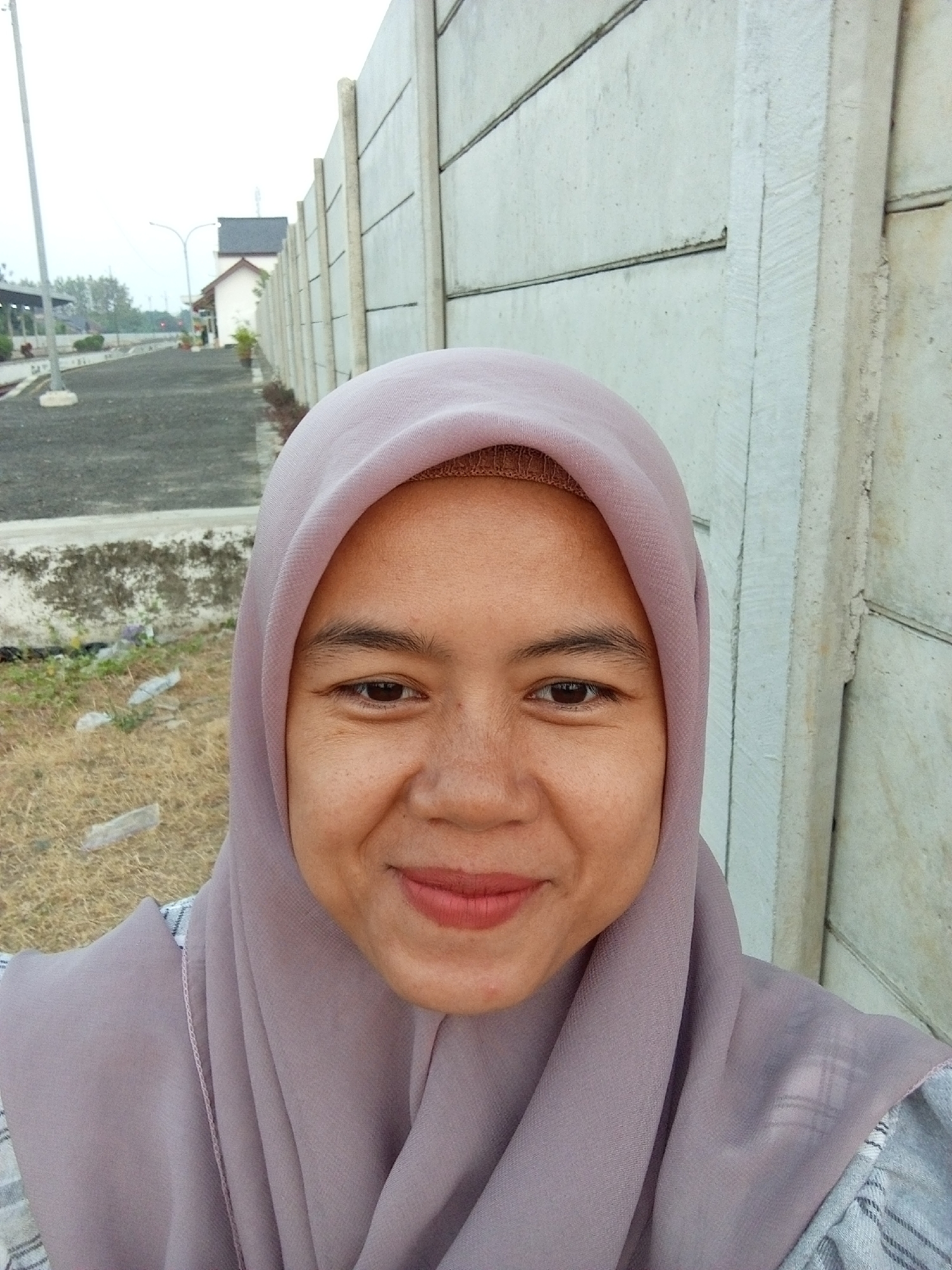 A medium shot captures a young Indonesian woman smiling at the camera, positioned in front of a light gray, concrete wall. She is wearing a lavender hijab, the fabric draped over her head and shoulders. Her face is illuminated, revealing freckles across her nose and cheeks, and she has light pink lipstick on her lips. To the left of the wall, a gravel path is visible, leading towards a small building with a dark roof and white walls, situated in a rural or suburban area. The background also shows sparse vegetation and a street lamp. The overall scene appears to be outdoors, with the concrete wall forming a stark backdrop.