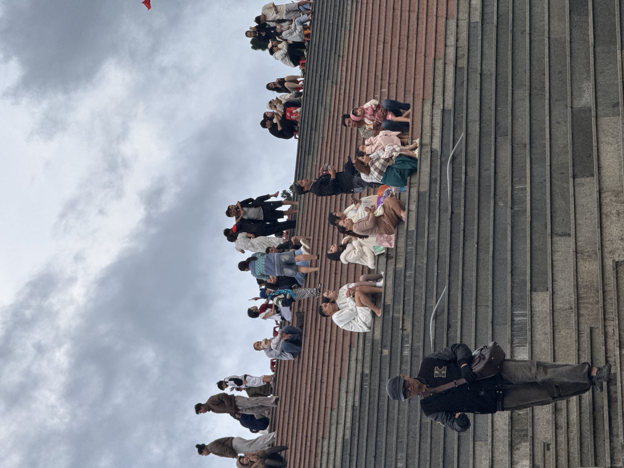 This is a photograph of the Dinh Ba Dai (former Emperor Bao Dai's summer palace) in Da Lat, Vietnam, showing a crowd of people gathered on wide, tiered stone steps leading up to the palace. The sky is overcast with dramatic clouds.
In the foreground, a man wearing a dark jacket with a yellow "K" emblem, dark pants, a cap, and glasses walks down the steps. He carries a brown satchel over his shoulder.
Further up the steps, various groups of people are seated and standing. On the lower of these upper tiers, a couple sits closely together, the woman wearing a white outfit and the man in a light-colored top and shorts. To their right, another woman in a white coat sits with a young child.
On a higher tier, a man in a black shirt stands and appears to be speaking or gesturing. A group of women and children are seated on the steps, some holding bags and one child wearing a pink hooded garment.
Higher still, more people are seated and conversing. On the topmost visible tier, several individuals are standing and sitting, observing the surroundings. A small red flag is visible in the far upper right corner of the frame, against the cloudy sky. The steps are made of reddish-brown stone on the upper section and gray stone on the lower section.