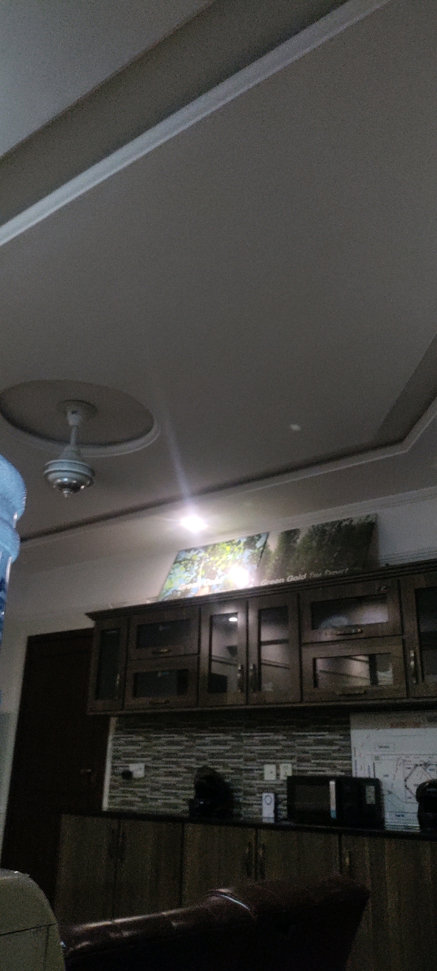 A low-angle, indoor photograph shows a ceiling with a recessed center featuring a decorative molding and a white ceiling fan with silver blades. To the right, a brightly lit framed artwork depicting a forest scene is mounted on the wall. Below the artwork, a dark wooden cabinet with glass-fronted doors spans the width of the wall. To the left of the cabinet, a dark brown door is visible. The background wall behind the cabinet is covered in a horizontal patterned tile. On the countertop above the lower cabinets, a microwave oven, a small white object, and a map are placed. The image is dimly lit, with light sources causing lens flare. The location is identified as Khana Dak, Pakistan.