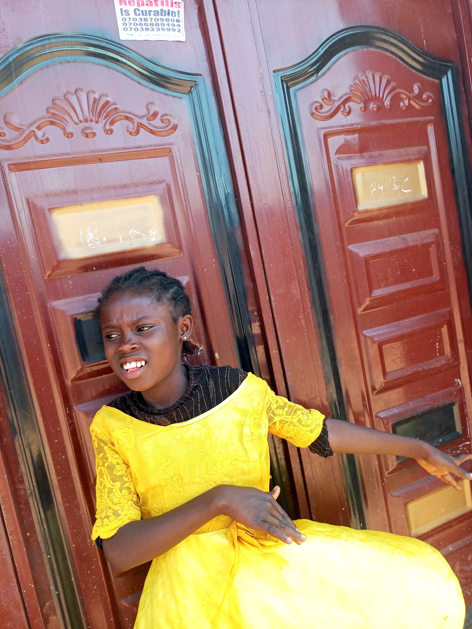 A young girl with dark skin and braided hair is standing in front of a brown, paneled door with ornate carvings and decorative moldings. She is wearing a bright yellow dress with lace sleeves and black trim. Her left hand is resting on her lap, while her right arm is extended to her right, with her hand reaching out towards the door. She has a slightly distressed expression on her face, with her mouth open and her brow furrowed. A partially visible sticker on the door in the upper left corner contains text in pink and blue, and appears to mention "Hepatitis" and several phone numbers. The scene is likely an outdoor setting, possibly in the city of Gombe, Nigeria, given the presence of the door and the visual style of the sticker, which is consistent with local advertisements in that region.