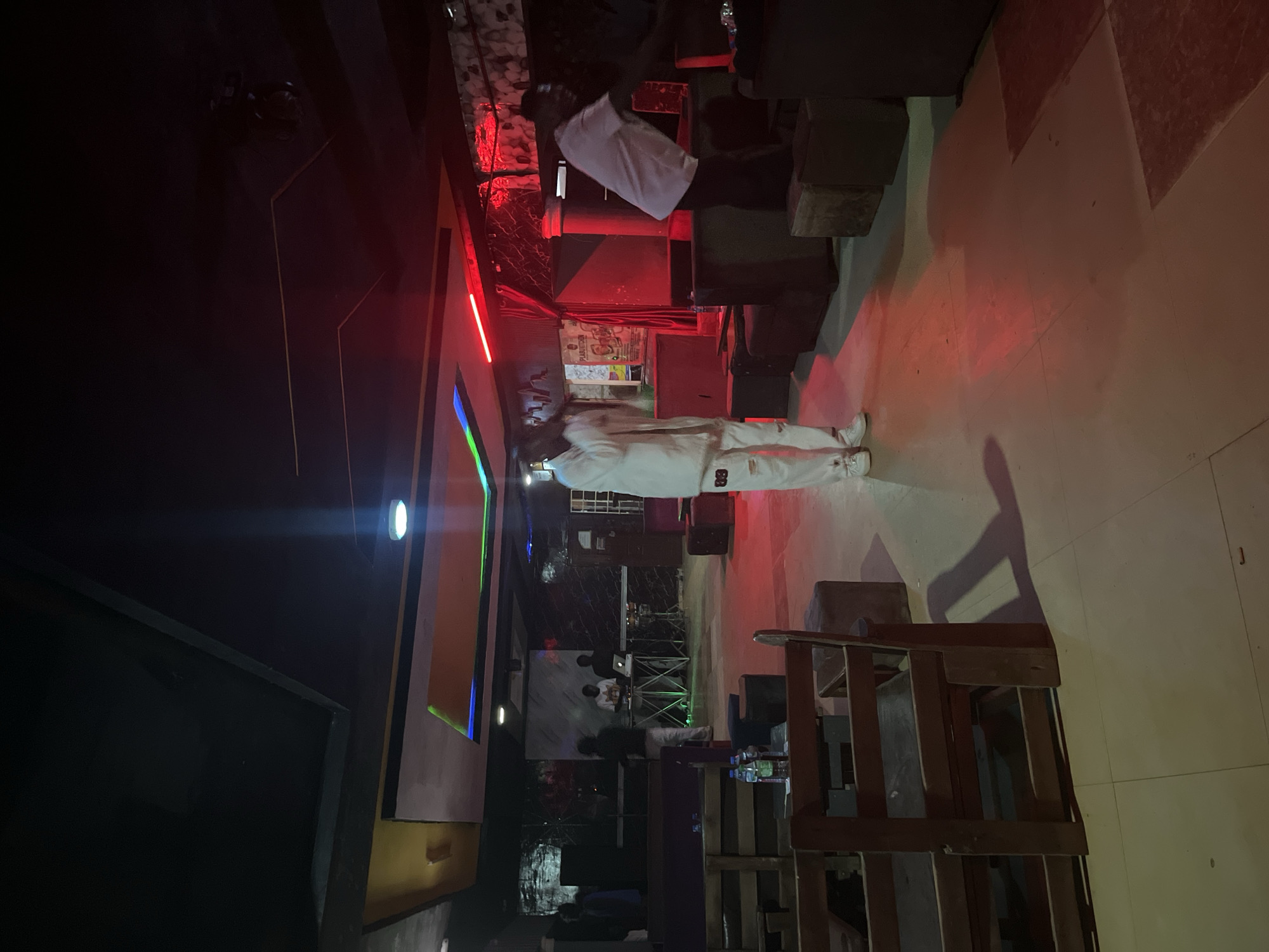 The image depicts an indoor scene within a venue in Durumi, Nigeria, likely a club or bar. The lighting is dim, with prominent red and blue neon accents on the ceiling and walls, creating a club-like atmosphere. In the foreground, wooden chairs and tables are sparsely arranged.
The central focus is on a person in the middle ground, wearing a white tracksuit with white sneakers, holding a microphone and appearing to perform or address an audience. To the right, another person, wearing a white t-shirt and dark pants, is partially visible, leaning forward as if interacting with equipment or an individual to their right, out of frame.
In the background, a DJ setup is visible with at least two individuals tending to it. Various seating arrangements, including couches and cubes, are scattered throughout the space. The floor is tiled. The overall impression is of a nightlife establishment during an event.