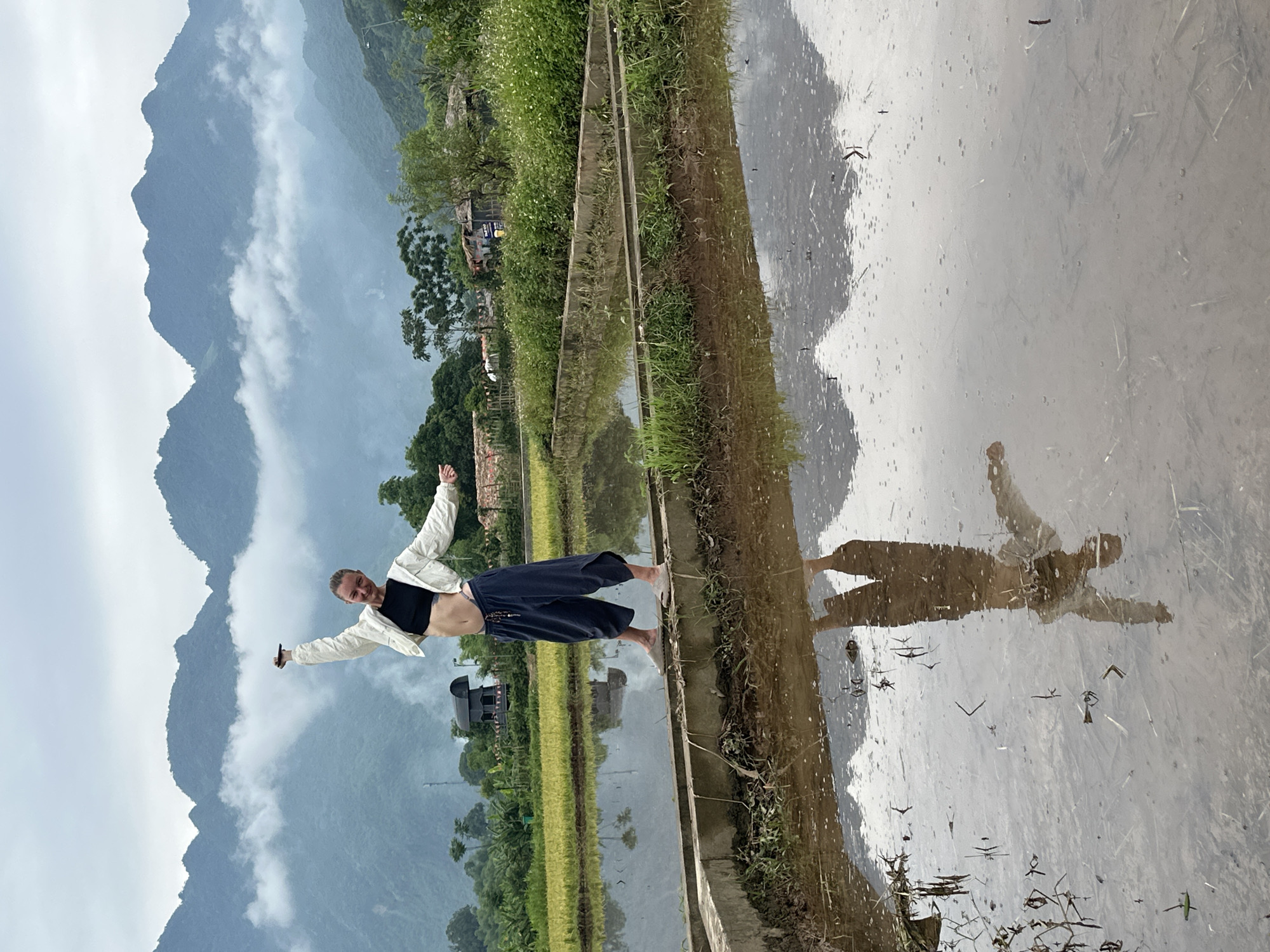 A young Caucasian woman with blonde hair pulled into a bun stands on a narrow concrete ledge dividing flooded rice paddies in Mai Chau, Vietnam. She is wearing a black crop top, a white jacket, and loose dark pants. Her arms are outstretched and her head is tilted back, as if in a moment of joy or celebration. In the background, lush green rice fields stretch towards mist-shrouded mountains. The water in the paddies reflects the sky and the woman, creating a mirror image. A few simple buildings and trees are visible in the mid-ground, adding to the rural atmosphere. The sky is overcast, with soft clouds hanging low over the mountains.