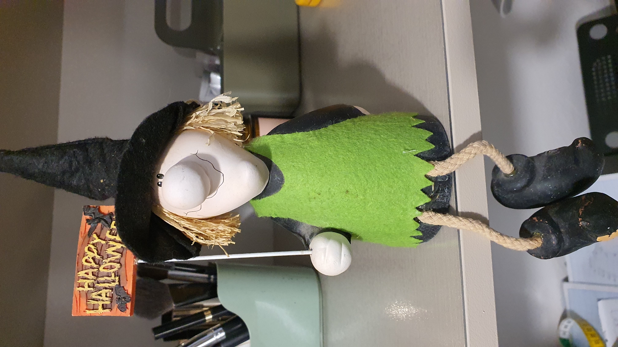This media file features a close-up, horizontal shot of a Halloween-themed decorative figurine. The figurine depicts a witch with a pointed black hat and a green, jagged-edged dress. She has a pale, round face with painted-on eyes and a nose. Her hair appears to be made of straw. In her left hand, she holds a small, orange sign with the words "HAPPY HALLOWEEN" written in yellow block letters, adorned with small black bat figures. The witch's arms are not visible, but her legs are depicted as rope-like material attached to black, shoe-like feet.
The figurine is positioned on a light-colored shelf or surface. Behind the witch, a blurred background suggests an indoor setting with a light-colored wall and some shelving units. To the right of the witch, a black electronic device with circular indentations is visible. Below the witch's feet, a set of makeup brushes or art tools are partially visible in a container. The overall impression is of a decorative item for a holiday celebration, likely placed on a shelf or desk. The lighting appears to be artificial, and the scene is well-lit, suggesting it was captured indoors during the day or evening. No specific time of day or weather conditions can be inferred. The location is identified as Krasnoyarsk, Russia.