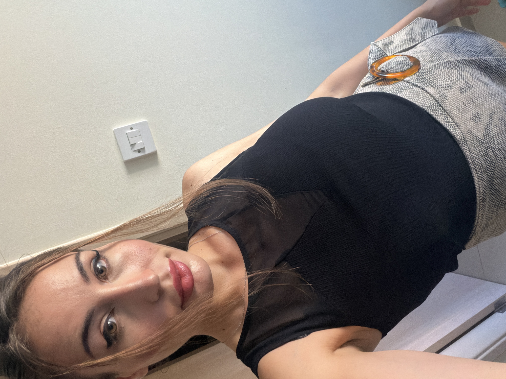 A woman is taking a selfie indoors. She is wearing a black sleeveless top with a sheer neckline and a patterned skirt with an orange ring detail. The background is a plain wall with a light switch. There is a doorway on the left side of the frame. The lighting suggests it is daytime. The location is identified as Tersefanou, Cyprus.