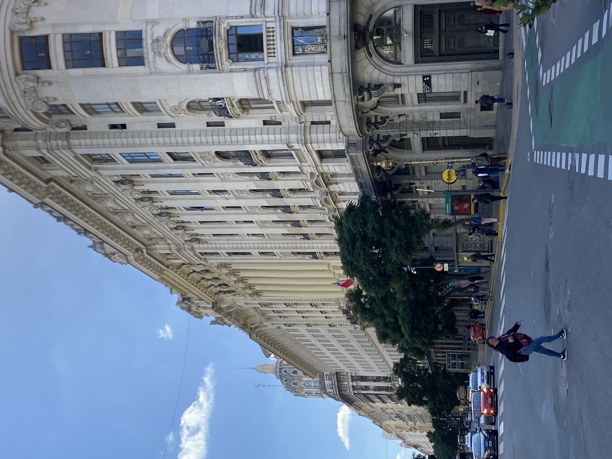 The image, currently displayed 90 degrees counter-clockwise from its natural orientation, presents a vibrant street scene in Buenos Aires, Argentina. The setting is daytime under a bright blue sky dotted with scattered white clouds.
To the left, a grand, multi-story building with classical architectural elements dominates the frame. Its facade is adorned with decorative cornices, columns, arched windows, and balconies featuring intricate ironwork. Further along the street, additional ornate buildings are visible, including one in the background topped with a distinctive dome and spire. Green trees line the sidewalk in front of these structures, and a small red and white flag hangs from one building.
The right side of the image features a paved street with distinct markings, including a green-painted bicycle lane and a white-striped crosswalk. Several cars are present on the street, some appearing to be in motion or parked. Pedestrians are scattered throughout the scene; some walk on the sidewalk, while others are captured in the act of crossing the street. In the immediate foreground, a person wearing a dark jacket, blue jeans, and carrying a bag is seen mid-stride, seemingly crossing the road.
Notable street furniture includes a prominent circular yellow sign with the text "Subte" (referring to the subway system) and "Catedral" written beneath it, indicating a station or nearby landmark. A 'no entry' or 'prohibited' traffic sign is also visible on a pole. The overall atmosphere suggests a bustling urban environment with historical architecture.