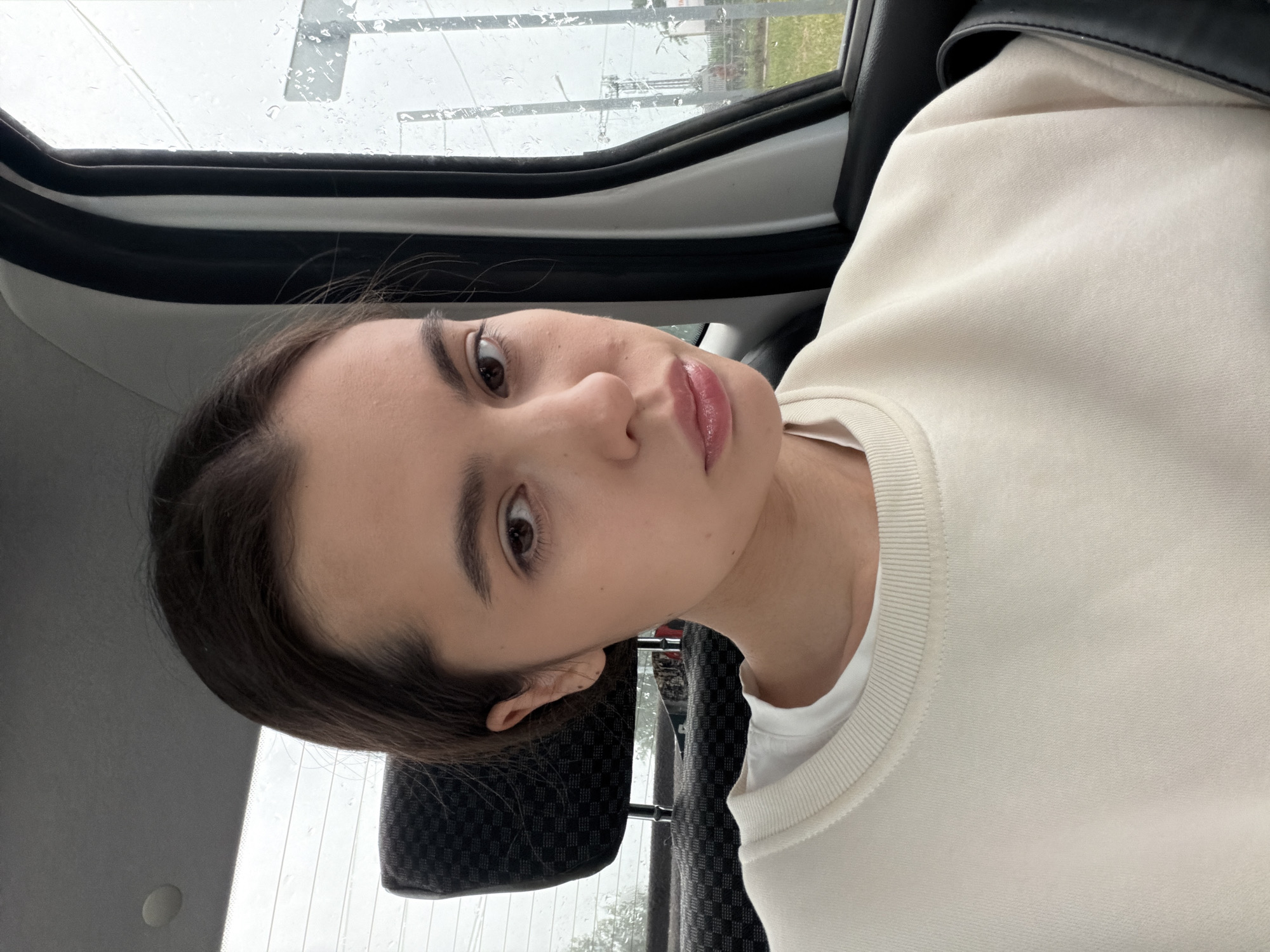 A young woman is captured in a close-up, portrait-style shot, with the image rotated 90 degrees clockwise. She gazes directly towards the viewer with a neutral, slightly contemplative expression. Her dark hair is pulled back, framing a clear face with prominent dark eyebrows, long eyelashes, and large, dark brown eyes. Her lips are full and appear to have a natural, glossy pink tint. She is wearing a light-colored, possibly cream or off-white, long-sleeved top, layered over a white t-shirt. A black strap, likely from a bag, is visible over her shoulder.
The setting is inside a car. A dark, checkered car seat headrest is visible behind her head and to her right. The car window frame runs along the upper part of the image, showcasing a window heavily covered in raindrops. Through the blurred, rain-streaked window, an outdoor scene is vaguely discernible, featuring green foliage and distant, indistinct structures under what appears to be an overcast sky. The visible raindrops indicate that it is raining heavily. The scene takes place during the daytime in Chaikovsky, Russia. There is no visible text in the image.