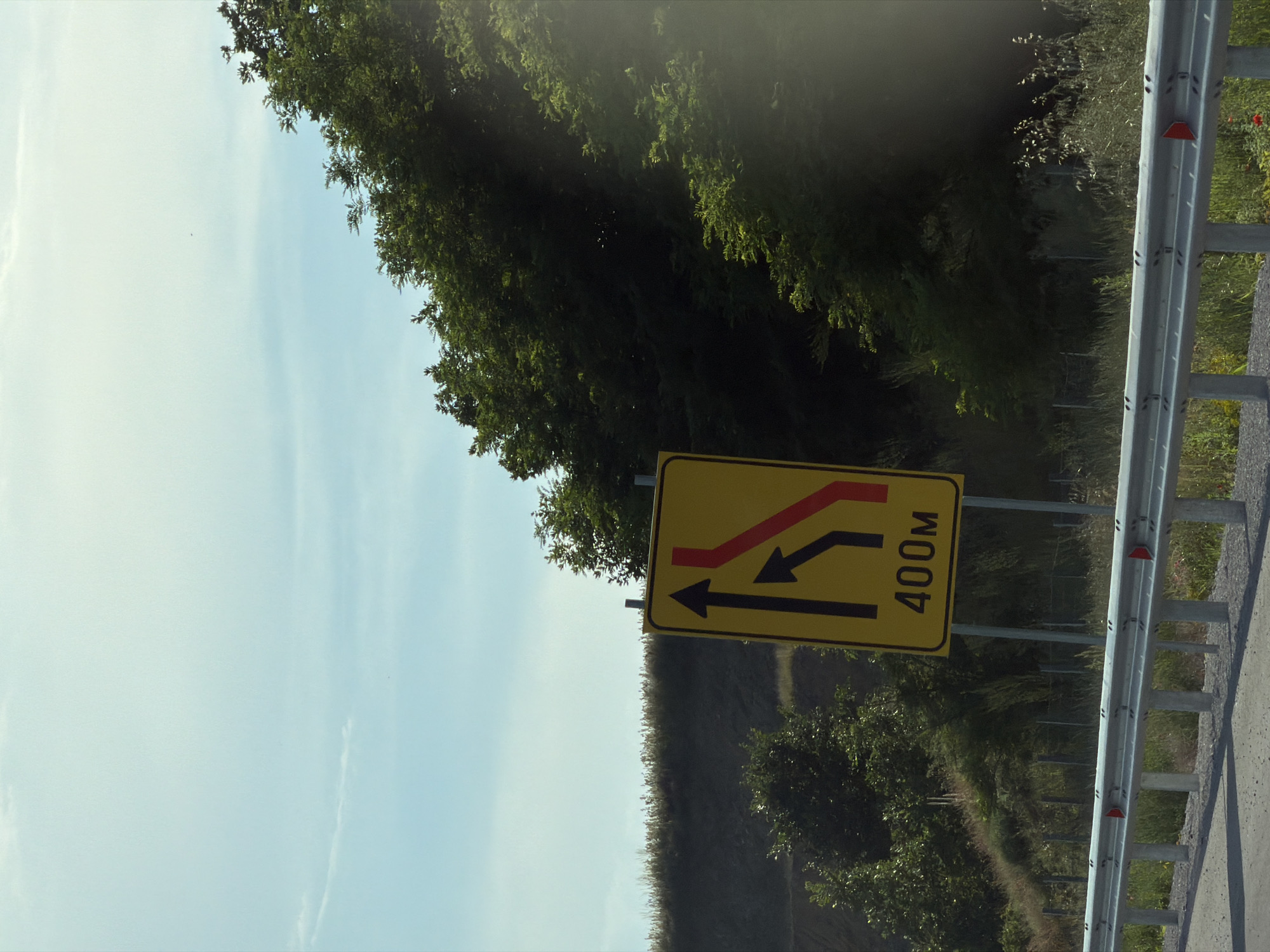 A yellow traffic sign with black and red arrows and the text "400m" is positioned on the right side of a road in Brestnitsa, Bulgaria. The sign indicates a lane reduction or merge ahead. The road is bordered by a metal guardrail. In the background, there is a grassy embankment covered with trees. The sky above is light blue with some wispy clouds. The scene appears to be a rural or roadside environment.