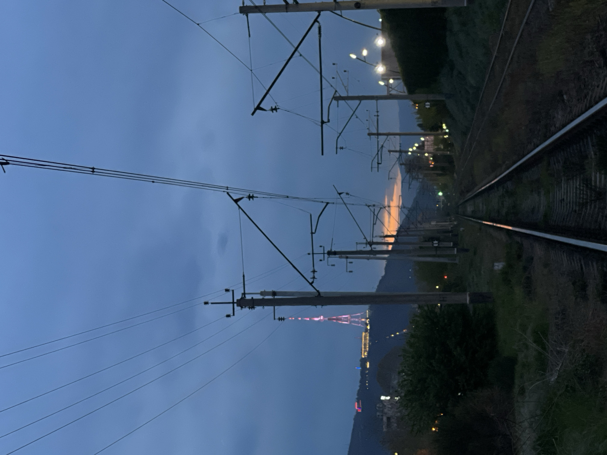 A vertical photograph, taken at dusk or early night, captures a railway line with prominent overhead power lines and poles. The railway tracks, likely a double-track system, stretch from the lower right towards the upper left of the frame. Numerous dark utility poles support a complex network of catenary wires, indicative of an electrified train route, silhouetted against a dark blue sky.
In the midground, dark green foliage and trees line the railway embankment, interspersed with streetlights that cast bright white and yellow glows. These artificial lights extend into the distant landscape, suggesting a city or town.
The background features a range of dark mountains. A distinctive and brightly illuminated television tower, glowing in red and pink hues, stands prominently on a distant hillside, identifying the location as Tbilisi, Georgia. Below and around the tower, a scattering of city lights dots the mountainous terrain. A subtle band of orange and yellow light is visible on the horizon, indicating the last remnants of sunset or civil twilight. The sky is largely a deep blue with some darker cloud formations. There are no visible people or discernible text in the image. The scene conveys a quiet, early evening atmosphere where urban infrastructure meets natural landscape.