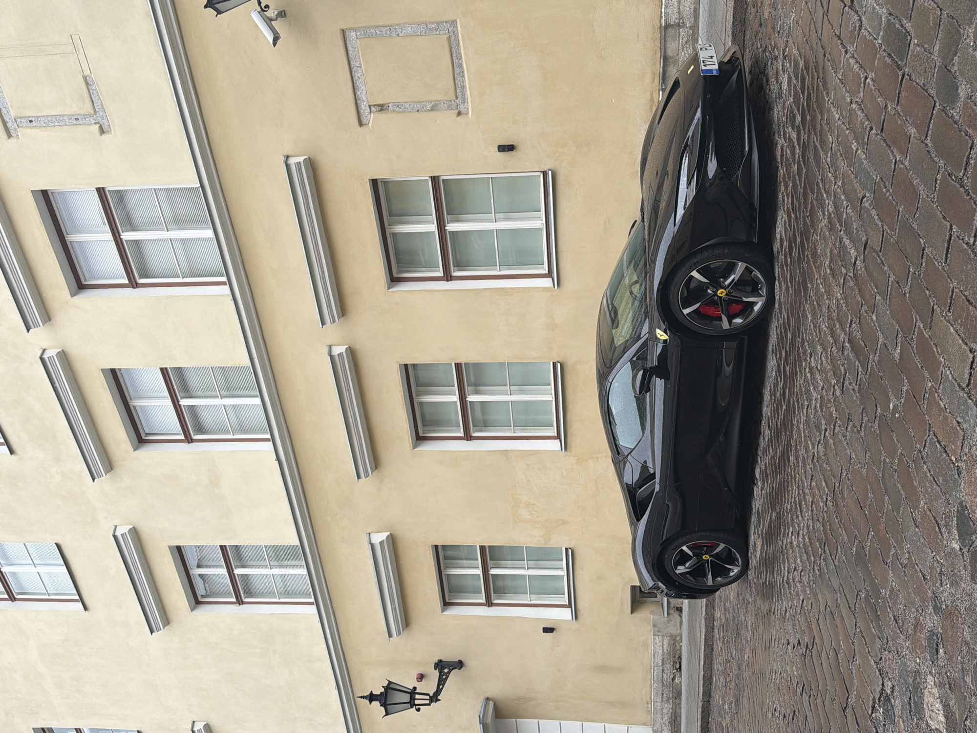 A black Ferrari sports car is parked on a wet cobblestone street in front of a pale yellow building in Tallinn, Estonia. The building has multiple windows with white blinds and decorative molding. A black lantern hangs from the wall to the left of the car, and a security camera is visible on the wall to the right. The car's license plate is visible and reads "174 PLS". The scene appears to be a quiet street with no people present.