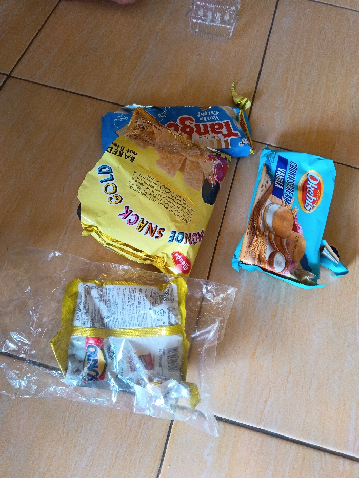 The image displays an assortment of snack packages laid out on a light brown tiled floor, indicative of an indoor setting in Cijeungjing, Indonesia. The lighting suggests it is daytime.

In the center, a crumpled yellow package is prominent, bearing the text "MONDE SNACK GOLD" and "BAKED not fried". Partially obscured above and to its right is a blue package for "Tango Vanilla Delight". To the right of the Monde snack, a light blue package of "Okebis" is visible, featuring an image of biscuits and labeled "COOKIES & CREAM Vanilla". In the lower-left corner, a clear plastic bag contains several smaller, individually wrapped items, with one red wrapper clearly showing the brand "UNO". A small, clear plastic container, possibly discarded packaging, is visible in the upper right portion of the image. The scene captures various packaged food items, likely snacks.