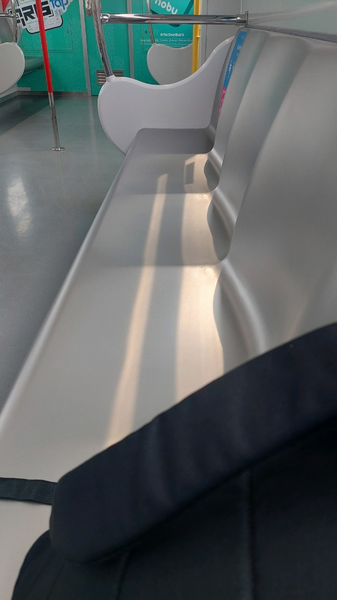 The image depicts the interior of a public transportation vehicle, likely a train or a bus, in North Jakarta, Indonesia. The seating is made of a light grey, smooth material, with a metallic sheen. The seats are arranged in rows, with the camera focused on the closest seat which appears to be empty. Sunlight is casting long shadows across the seats, suggesting it is either morning or late afternoon. In the background, teal-colored panels with white and green graphics are visible, some of which appear to be advertisements or informational signs. A red pole is also present on the left side of the frame, a common feature in public transport for passengers to hold onto. A dark-colored bag is partially visible in the foreground, obscuring a portion of the seat. The environment appears clean and well-maintained.