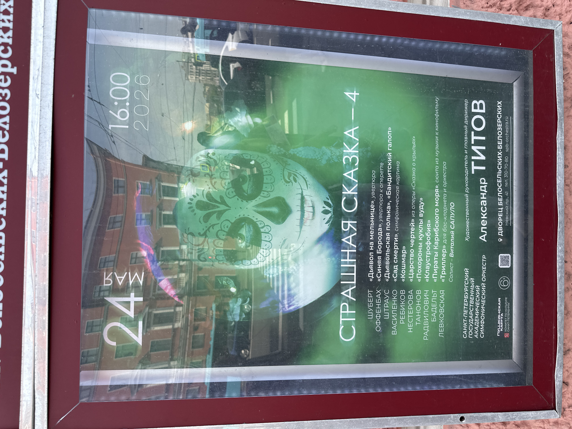The image is a close-up of a poster in a red frame advertising a performance of "A Scary Tale - 4" ("Страшная Сказка - 4") by the Saint Petersburg State Academic Symphony Orchestra, led by Alexander Titov. The poster lists a program of classical music pieces including works by Schubert, Offenbach, Strauss, Vasilenko, Rebikov, Nesterova, Tanonov, Radvilovich, Badelt, and Levkovskaya. The performance is scheduled for May 24, 2026 at 16:00 at the Beloselsky-Belozersky Palace, located at 41 Nevsky Prospekt, in Saint Petersburg, Russia. A stylized skull image forms the backdrop of the poster. Reflections of buildings are visible on the surface of the poster.