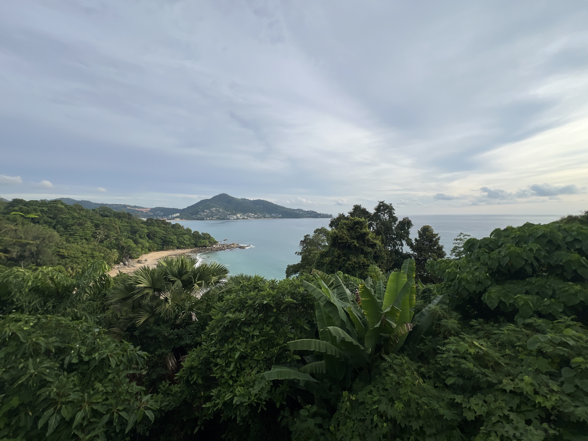 This image captures a serene coastal landscape in Ban Bang Thao, Thailand. The scene is dominated by lush, green tropical foliage in the foreground, with large, broad leaves and palm fronds suggesting a dense jungle environment. Beyond the dense vegetation, a curved, sandy beach is visible, bordered by the tranquil, turquoise waters of the ocean. Rocky outcrops are scattered along the coastline, adding texture to the scene.
In the distance, a verdant, hilly island rises from the sea, its slopes covered in trees and dotted with buildings that indicate a populated area. The sea stretches out to the horizon, meeting a sky filled with diffused light and scattered clouds. The overall impression is one of natural beauty and peacefulness, characteristic of a tropical paradise.
The lighting suggests it might be late afternoon or early morning, with the soft, overcast sky indicating a potentially cloudy or hazy day. There are no people or specific objects of human activity visible in the foreground or on the beach, emphasizing the natural environment. The scene evokes a sense of calm and relaxation. No visible text is present in the image.