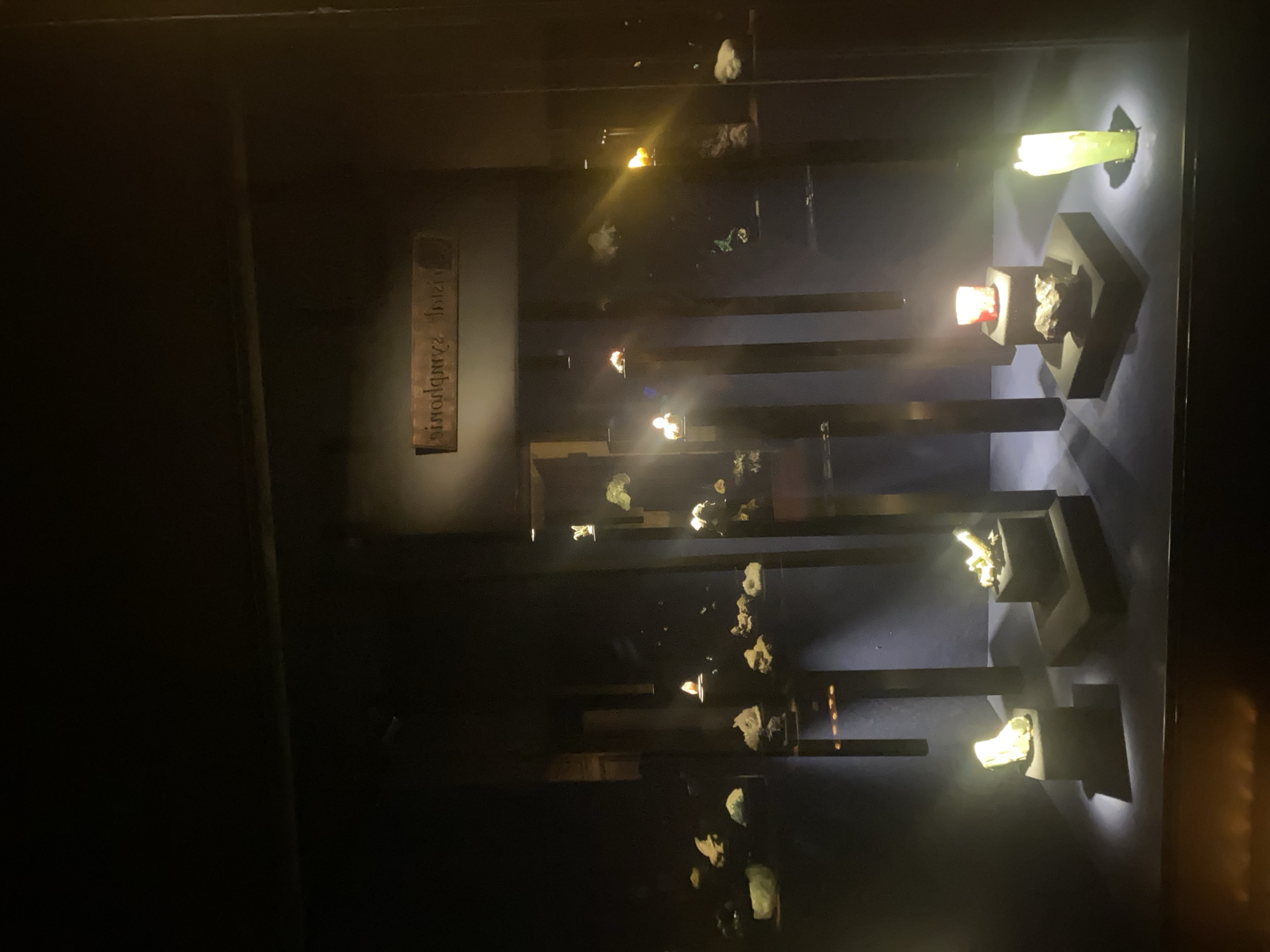 The image depicts a display case of minerals, likely in a museum or exhibit in Grenoble, France, as indicated by the visible text on a sign "Musée Grenoble". The objects are lit from above, highlighting the various rock and mineral samples. The display consists of vertical black bars with platforms holding the minerals. Some minerals are translucent and illuminated by backlighting. The overall scene is dimly lit, focusing the attention on the displayed items.
