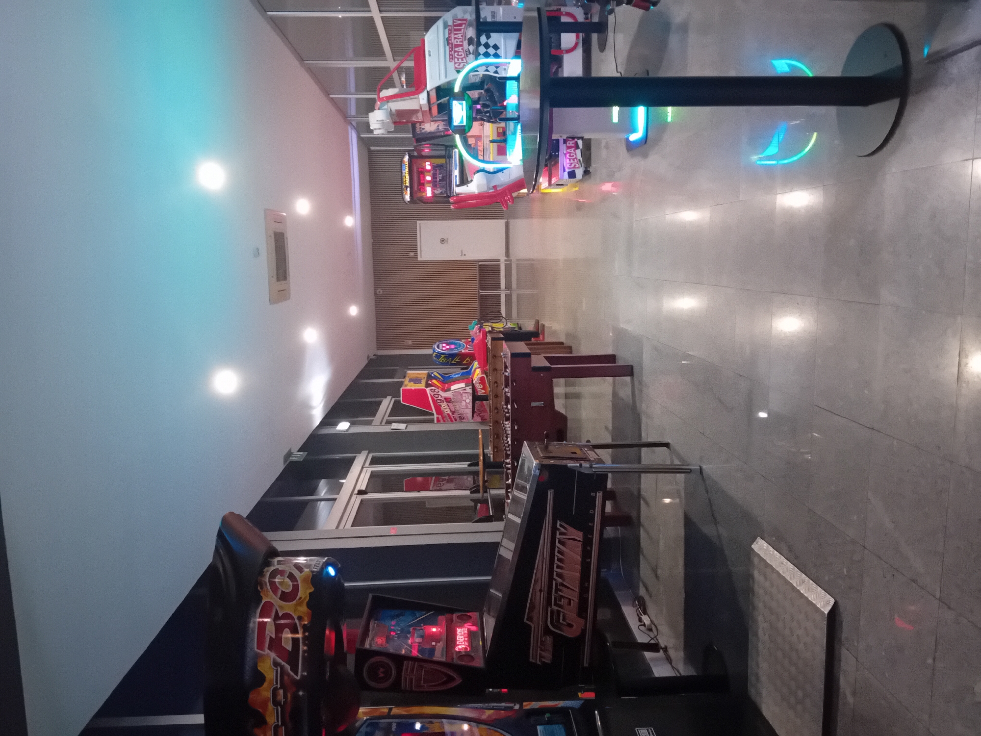 This indoor shot shows a hallway or arcade, likely in Figueira da Foz, Portugal. Several arcade game machines are visible, including racing games like Sega Rally. A black table with neon blue lights is also present, and the floor is a light grey marble. The ceiling has recessed lighting and a ventilation grill. The walls are light in color. No people are visible.
