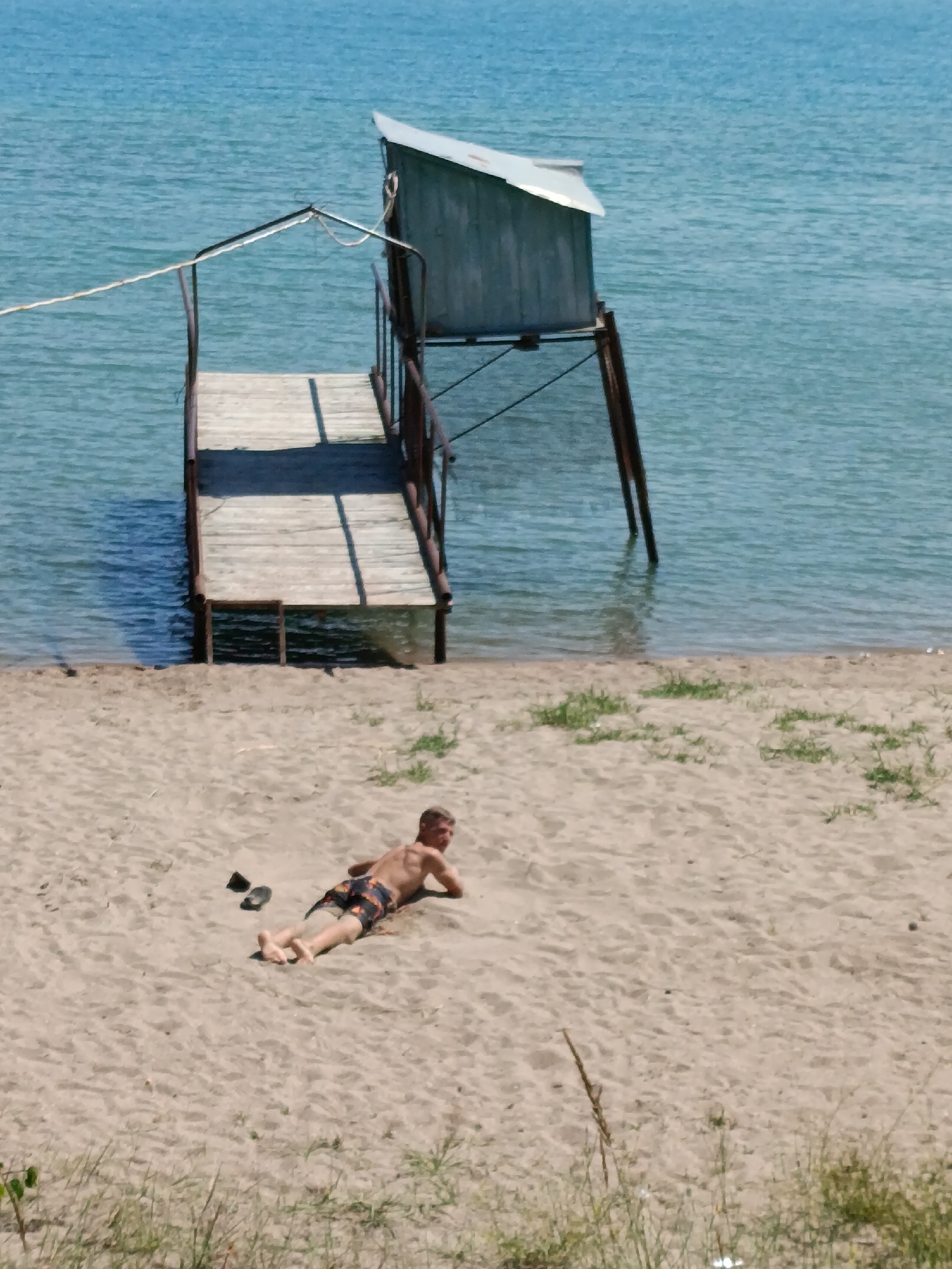 An elevated wooden pier extends from a sandy beach into the calm, blue waters of Issyk-Kul lake in Kyzyl-Suu, Kyrgyzstan. The pier has a rustic, weathered appearance, with metal railings and a small, wooden structure at its end, possibly a changing room or storage shed, situated on stilts. To the right of the pier, a young man is lying prone on the sand, facing away from the water and towards the left side of the frame. He is wearing patterned swim shorts and appears to be sunbathing. A pair of dark-colored flip-flops lies on the sand next to him. The beach is composed of fine, light-colored sand with sparse patches of dry grass and vegetation. The water is clear, with visible ripples on the surface. The overall scene depicts a serene day at the lake.