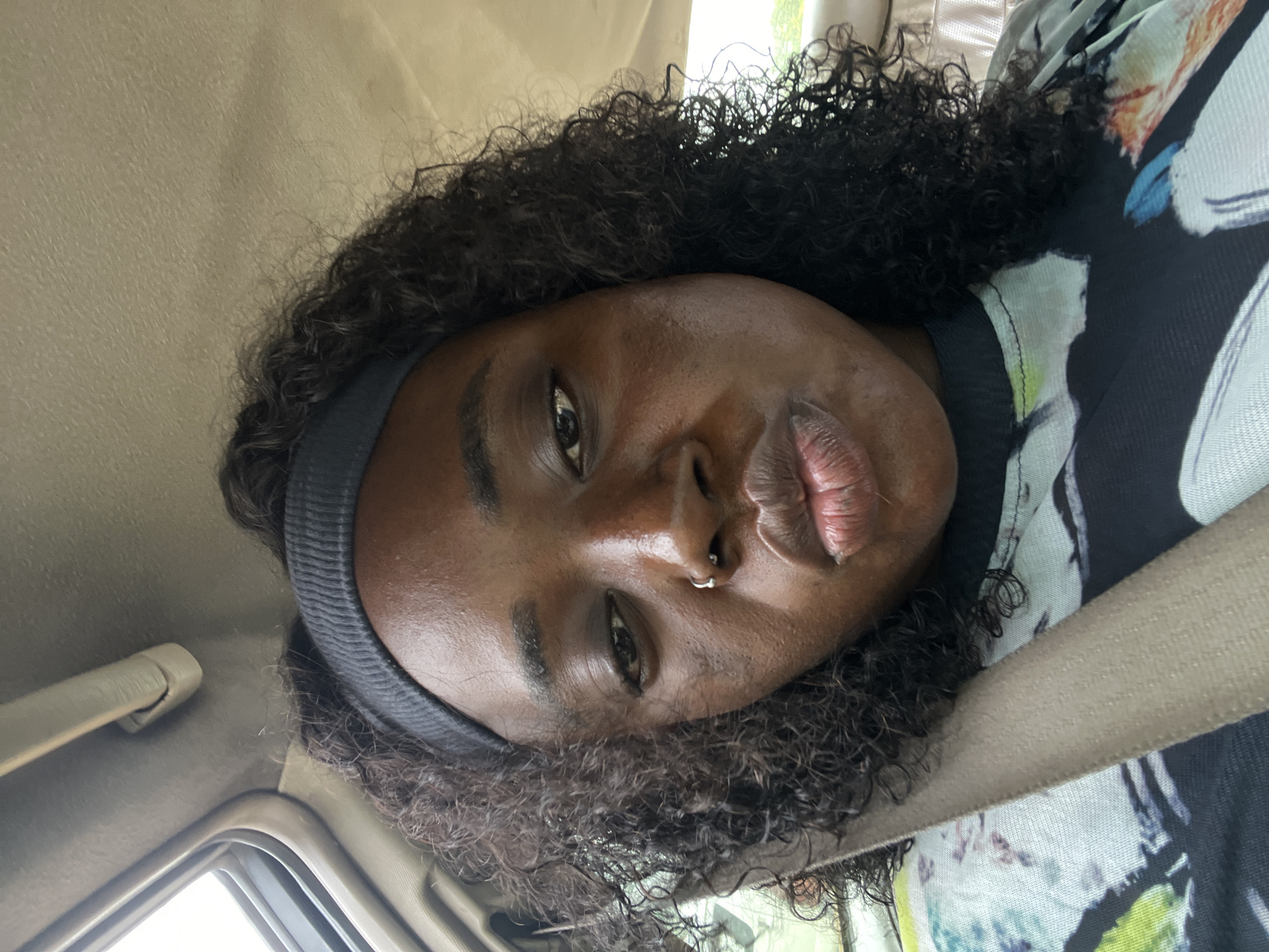 A medium shot captures a Black woman in a car, looking down at the camera with her lips puckered. She is wearing a black headband and a t-shirt with a colorful abstract print. A beige seatbelt is visible across her chest. The car's interior, including the headliner and sunroof, is visible in the background. The lighting suggests it is daytime. The woman has dark curly hair and a nose ring.