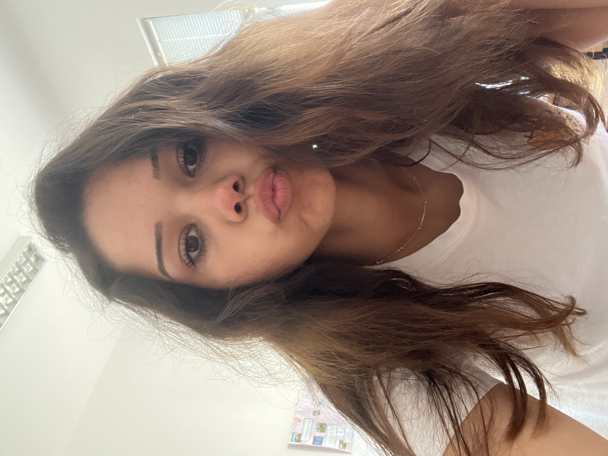 A young woman is featured in a selfie, captured from a slightly upward angle and oriented with her head on the right side of the frame. She has long, wavy brown hair with lighter highlights, which cascades around her face and shoulders. Her expression is playful, with her lips puckered into a pout and her eyes wide open, looking directly at the camera. Her eyelashes and eyebrows are prominent.
She is wearing a simple white t-shirt, and a delicate, gold-toned beaded necklace is visible around her neck. Her skin appears to have a warm undertone.
The background primarily consists of a plain, light-colored wall, likely white or off-white. In the upper left portion of the image, part of a white ventilation grate or a similar ceiling fixture is visible. In the lower right corner, a small section of what appears to be a bulletin board or display is seen, adorned with several small, indistinct pictures or notes. The scene is brightly and evenly lit, suggesting an indoor setting. No discernible text is visible in the image.