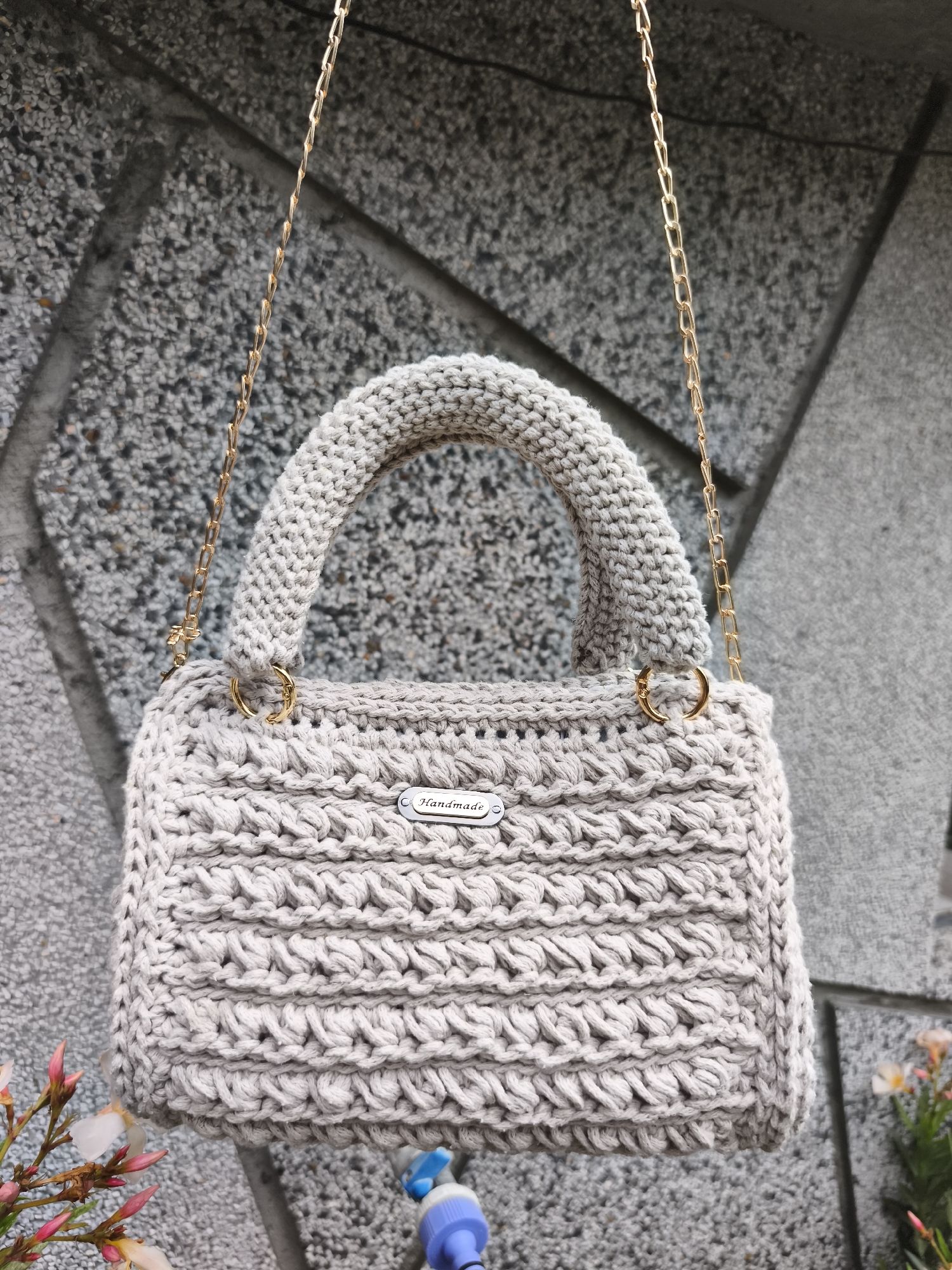 A crocheted handbag with a light beige color and gold chain straps hangs against a textured gray wall. The bag features a distinct woven pattern on its front and sides, and a small silver tag with the word "Handmade" is attached to the front. A padded handle, also crocheted, is connected to the bag by gold O-rings. The background wall appears to be made of stone or concrete with a rough, speckled surface. In the lower left corner, a few delicate pink flowers with green stems are visible. At the bottom center, a portion of a blue and white valve mechanism is peeking into the frame. The image is taken outdoors, with the light suggesting it is daytime. The location is identified as Marinka, Bulgaria.